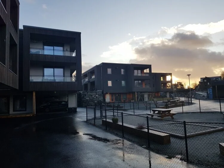 Moderne leilighetsbygg og lekeplass ved solnedgang, med asfaltert område og gjerder.
