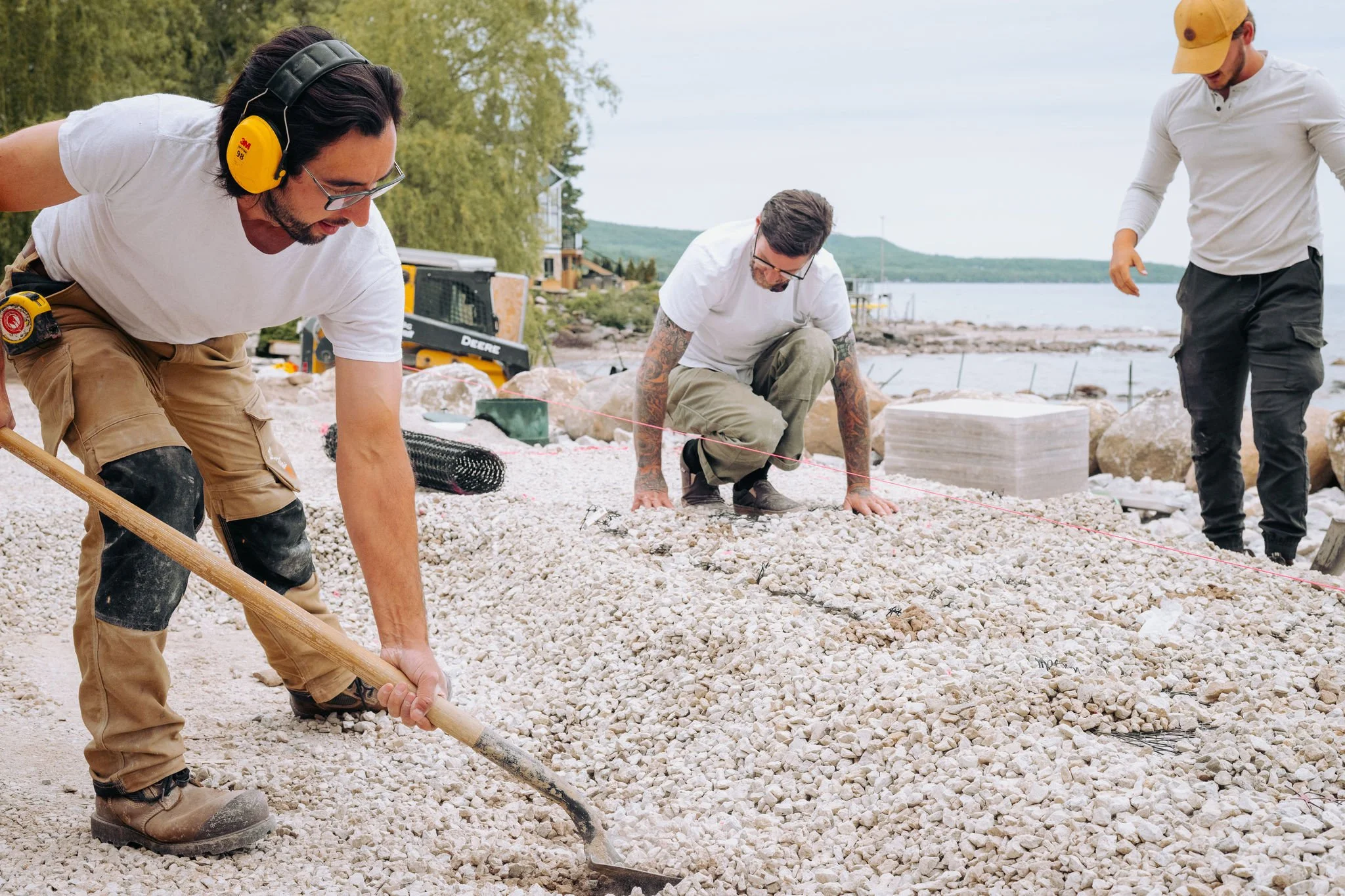 Three men working on a shoreline construction project, leveling and spreading gravel with tools, near a body of water and trees in the background.