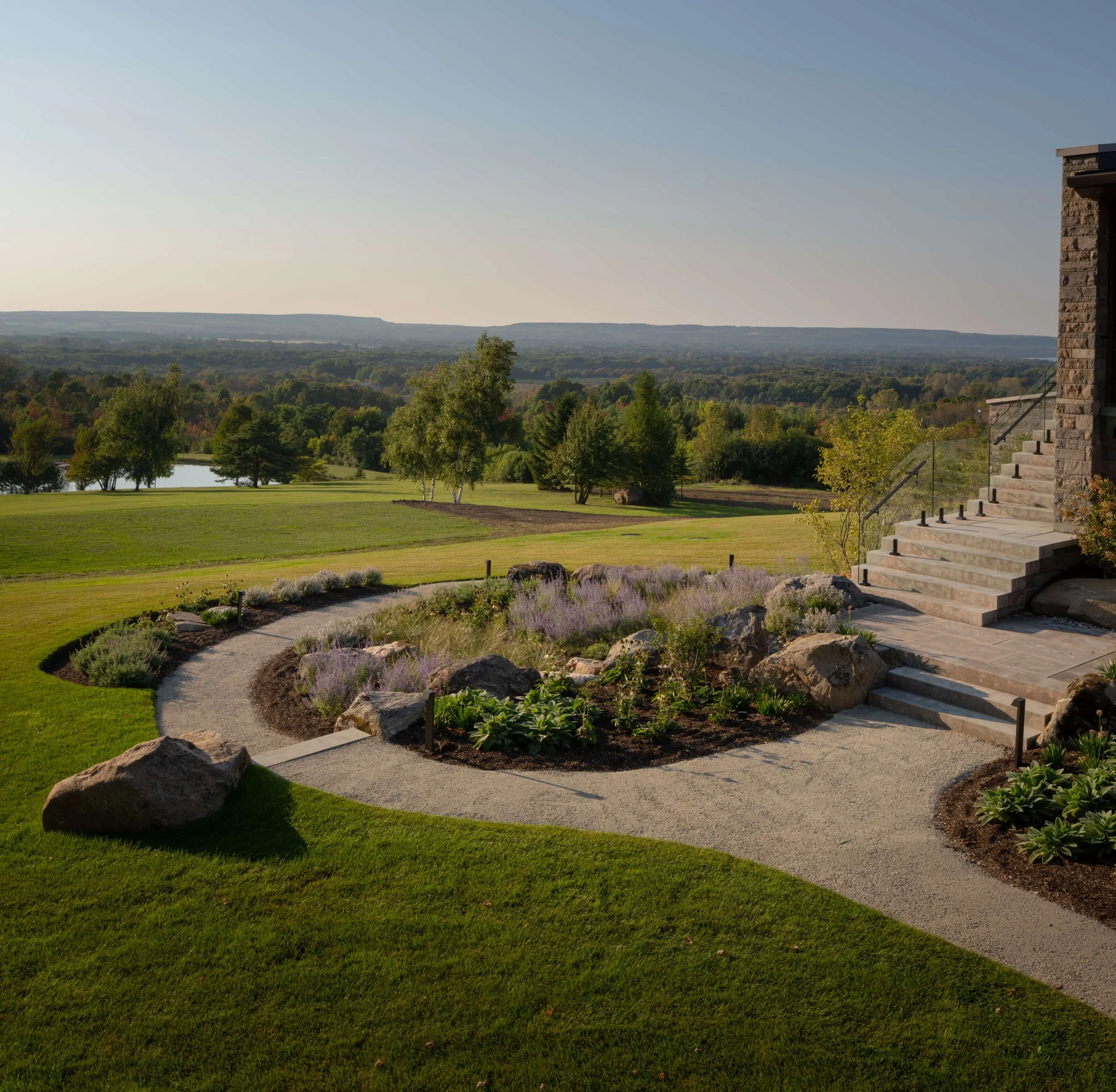 A landscaped yard with a curved gravel pathway, rocks, and garden plants next to a house with stone steps leading to a patio, overlooking a large green field and distant trees under a clear sky.