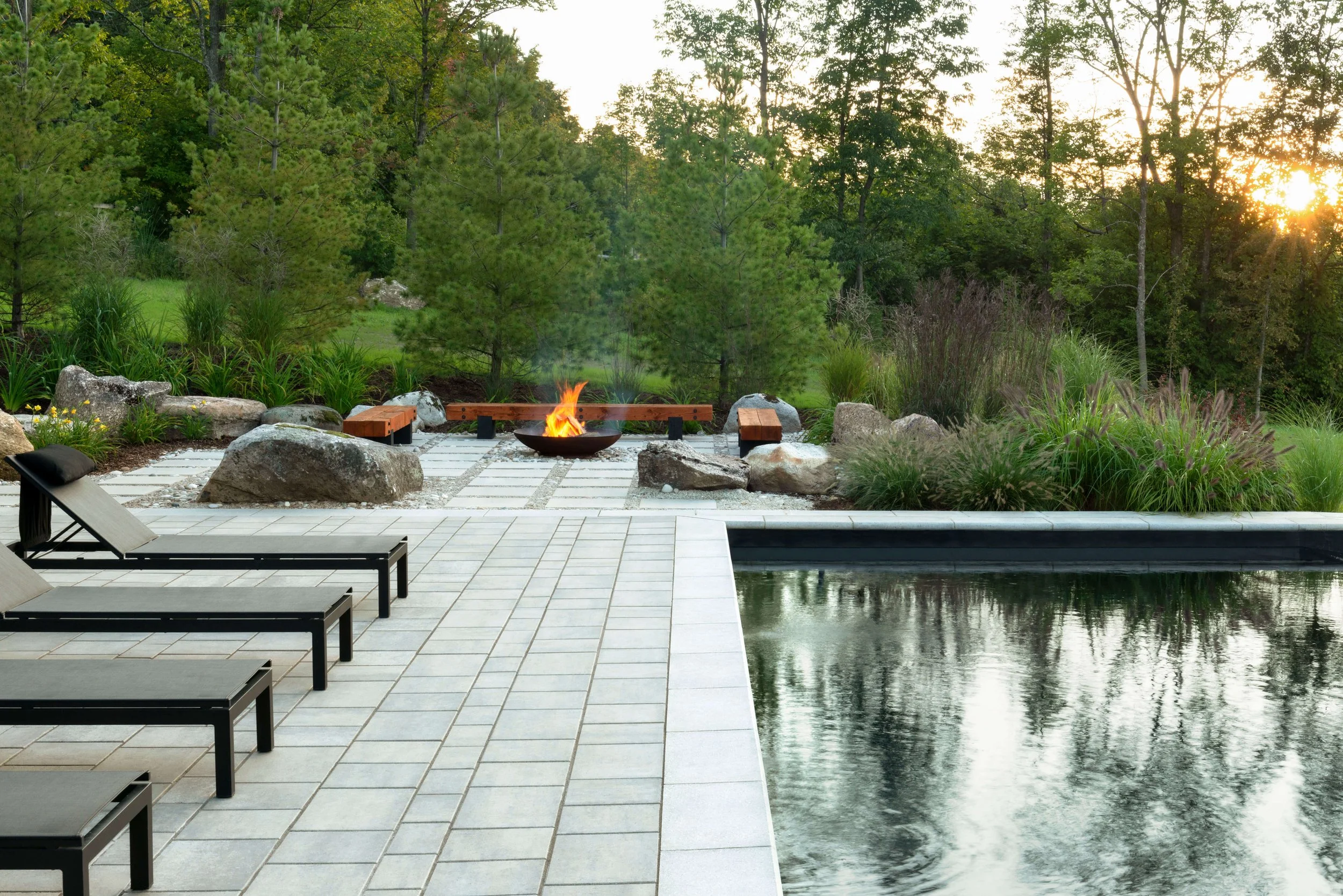Outdoor patio with a pool, lounge chairs, a fire pit, and surrounding greenery with trees and plants at sunset.