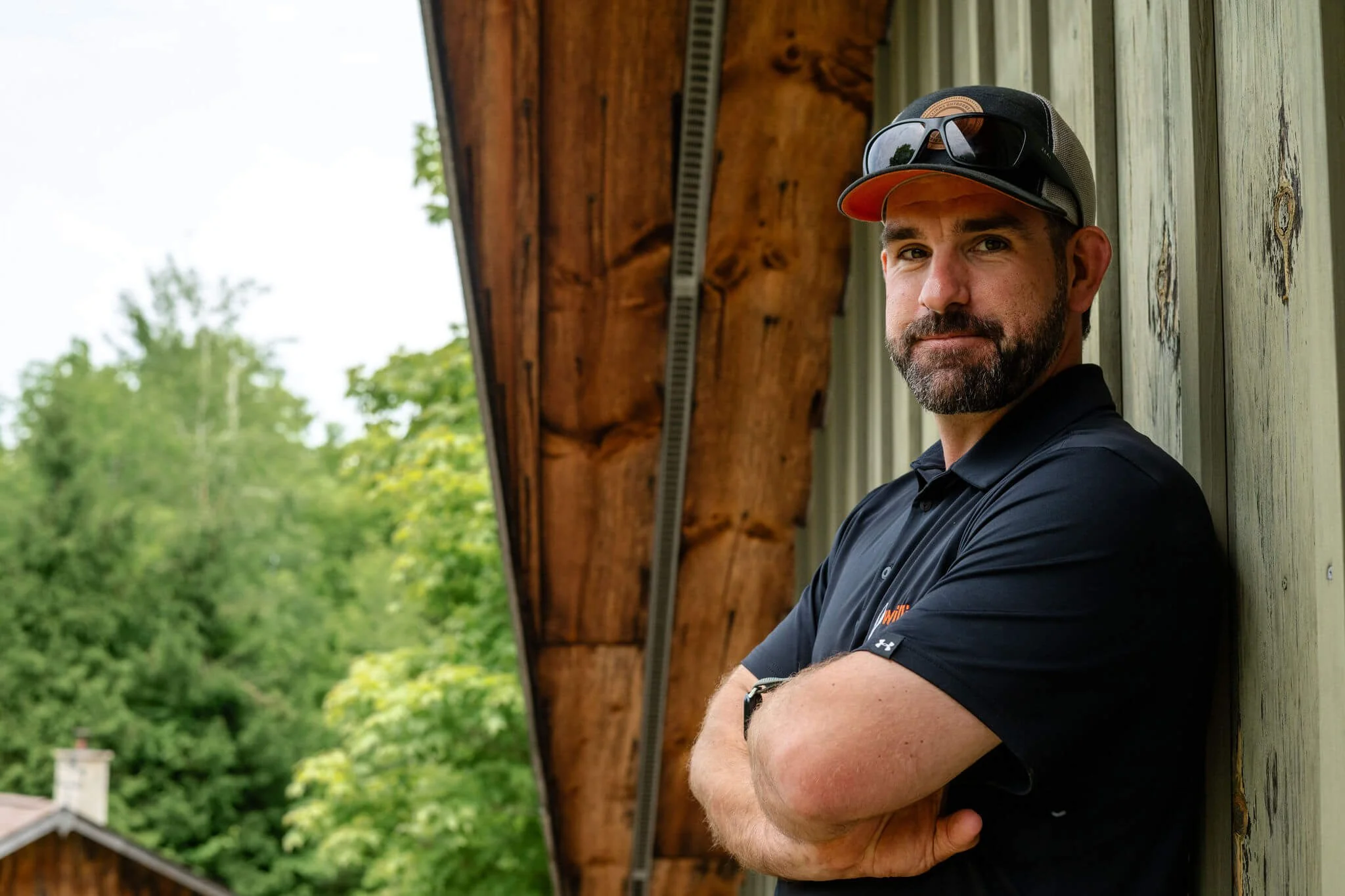 A man with a beard wearing a black polo shirt, sunglasses, and a baseball cap. He is standing outdoors with his arms crossed, leaning against a wooden wall with trees in the background.