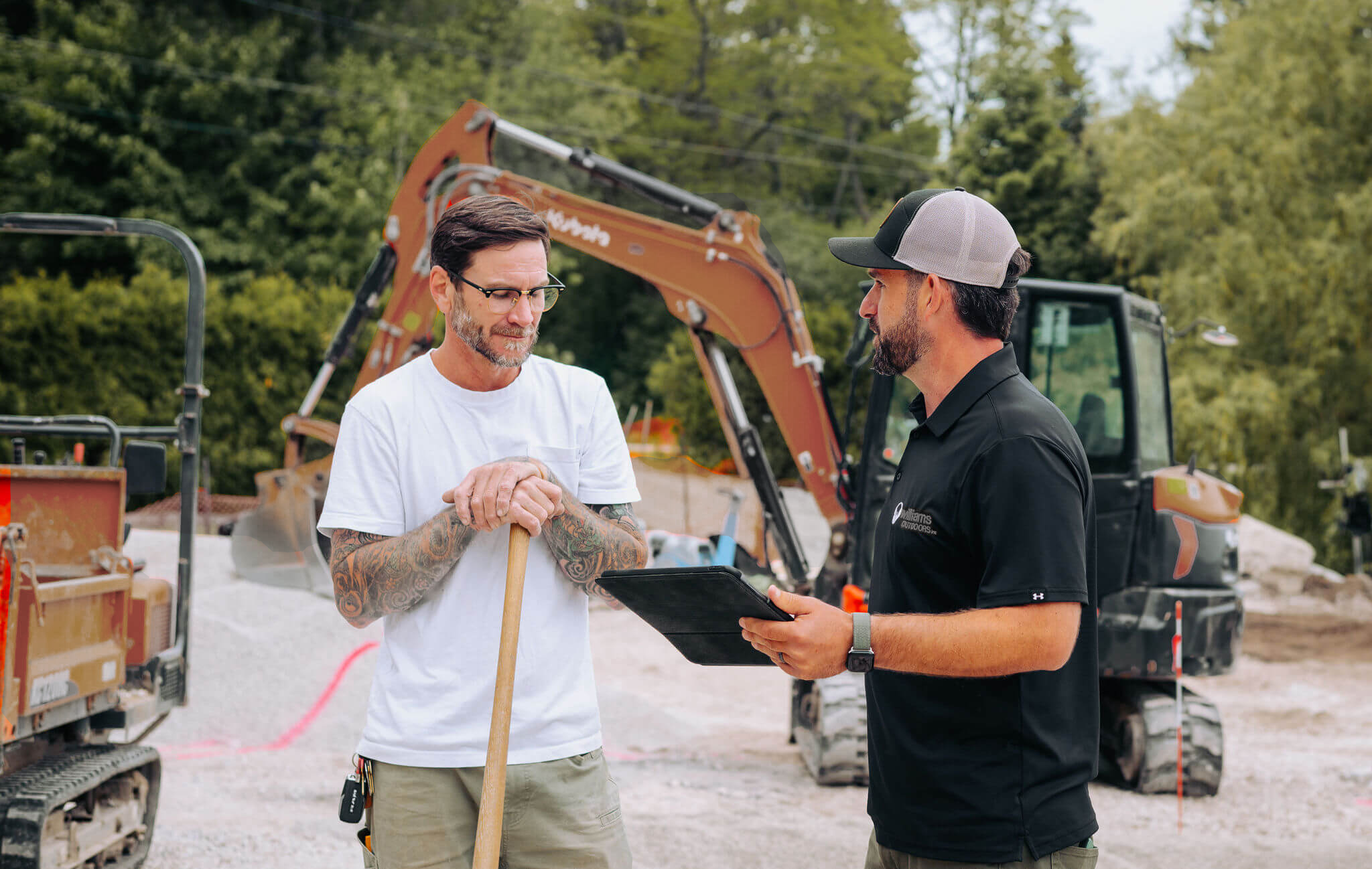 Two men having a conversation at a construction site with a backhoe and construction equipment in the background. One man holds a shovel, and the other has a tablet.