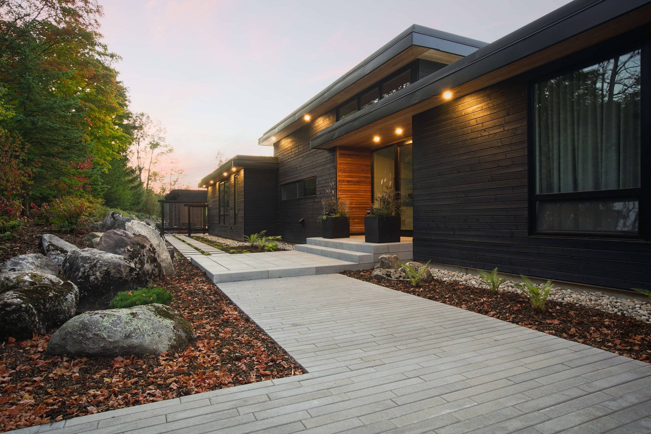 Modern black house with wooden accents, large windows, and outdoor lighting, featuring a paved walkway, rocks, small plants, and trees in a natural setting during sunset.