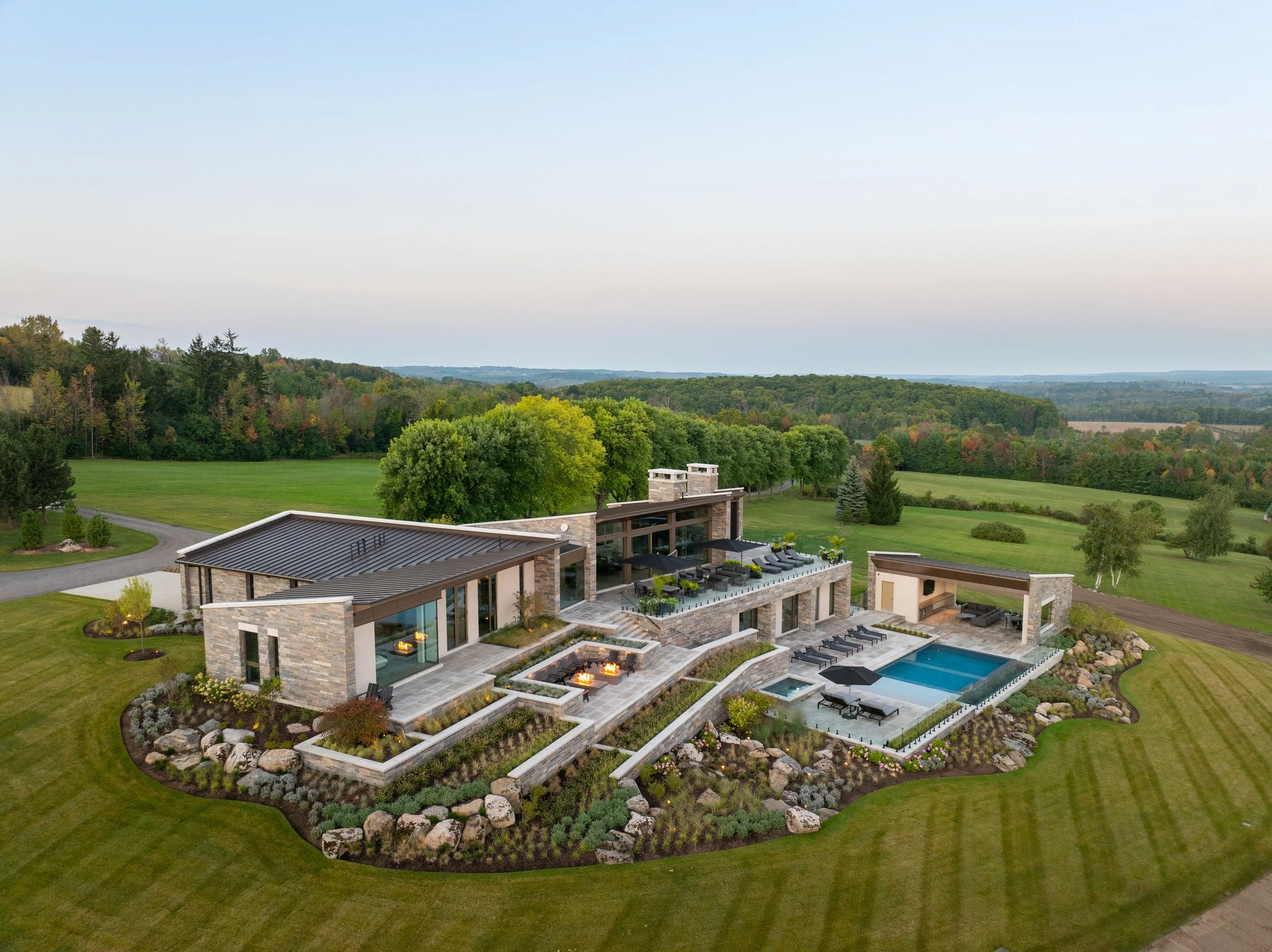 A modern house with a backyard pool and multiple fire pits, surrounded by a well-manicured lawn and landscape, with trees and rolling hills in the background.
