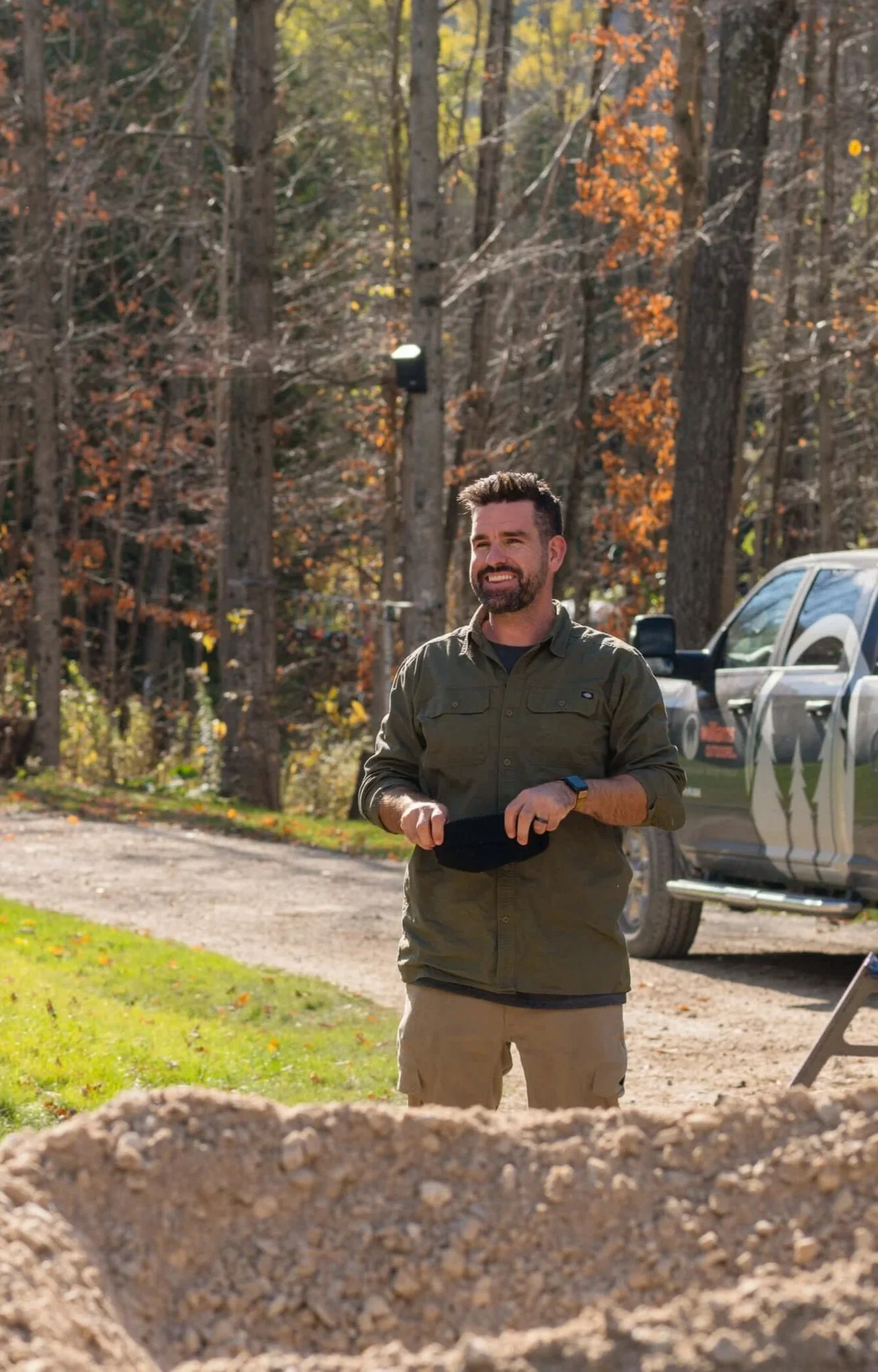 A man stands outdoors near a forested area with autumn leaves, holding a black cap and smiling, with a pickup truck in the background.