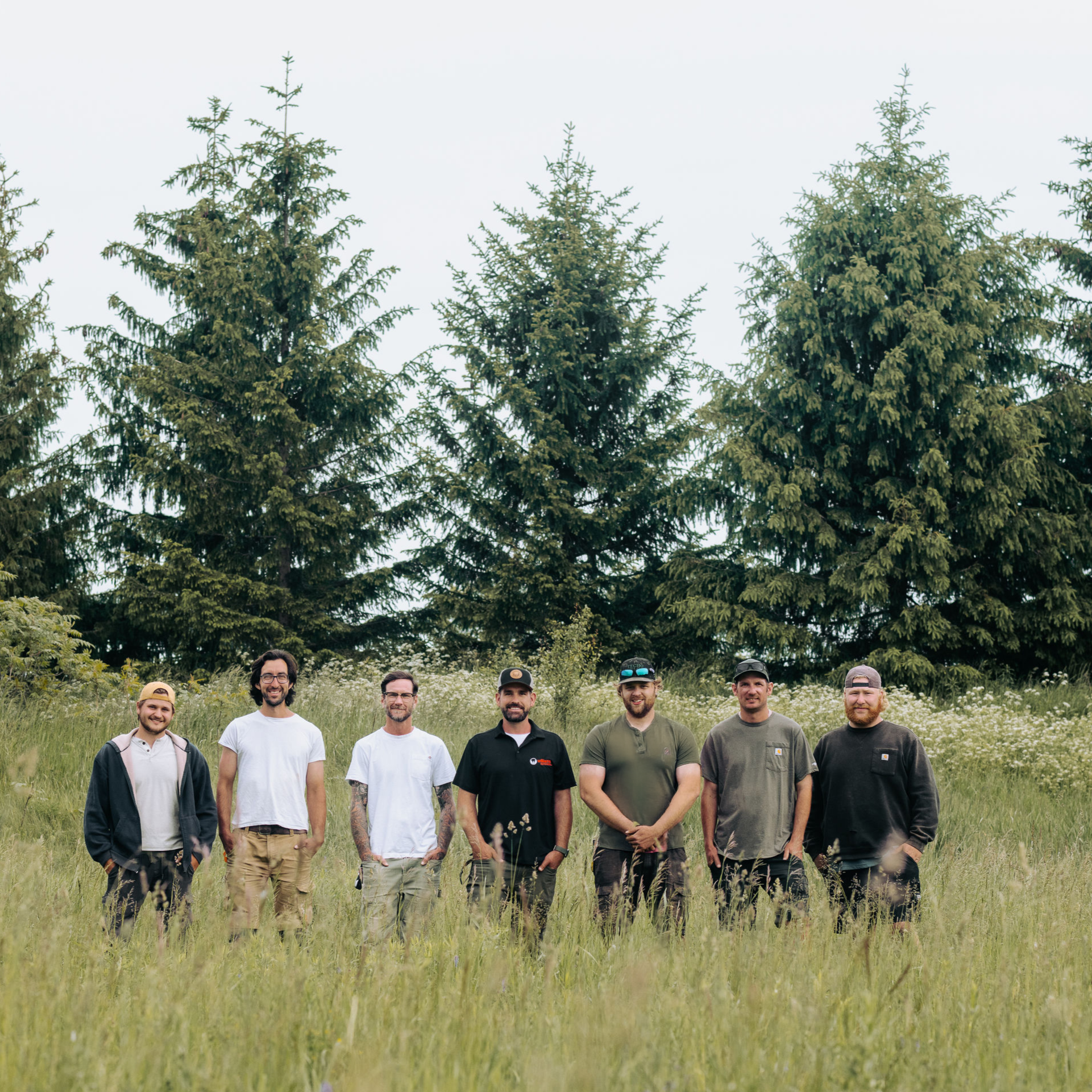 Group of seven men standing outdoors in a grassy field with tall evergreen trees in the background.
