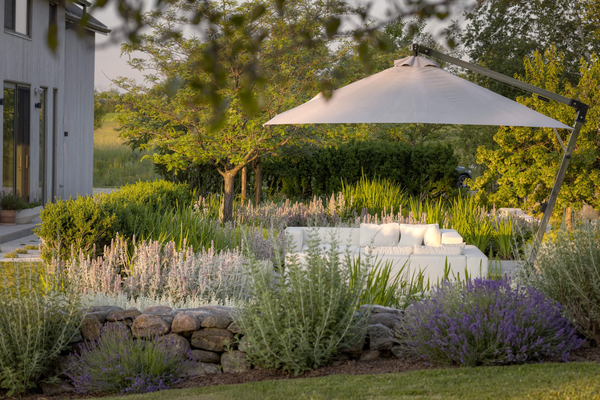 A white outdoor sofa under a large white patio umbrella in a lush garden with flowering plants and trees during sunset.
