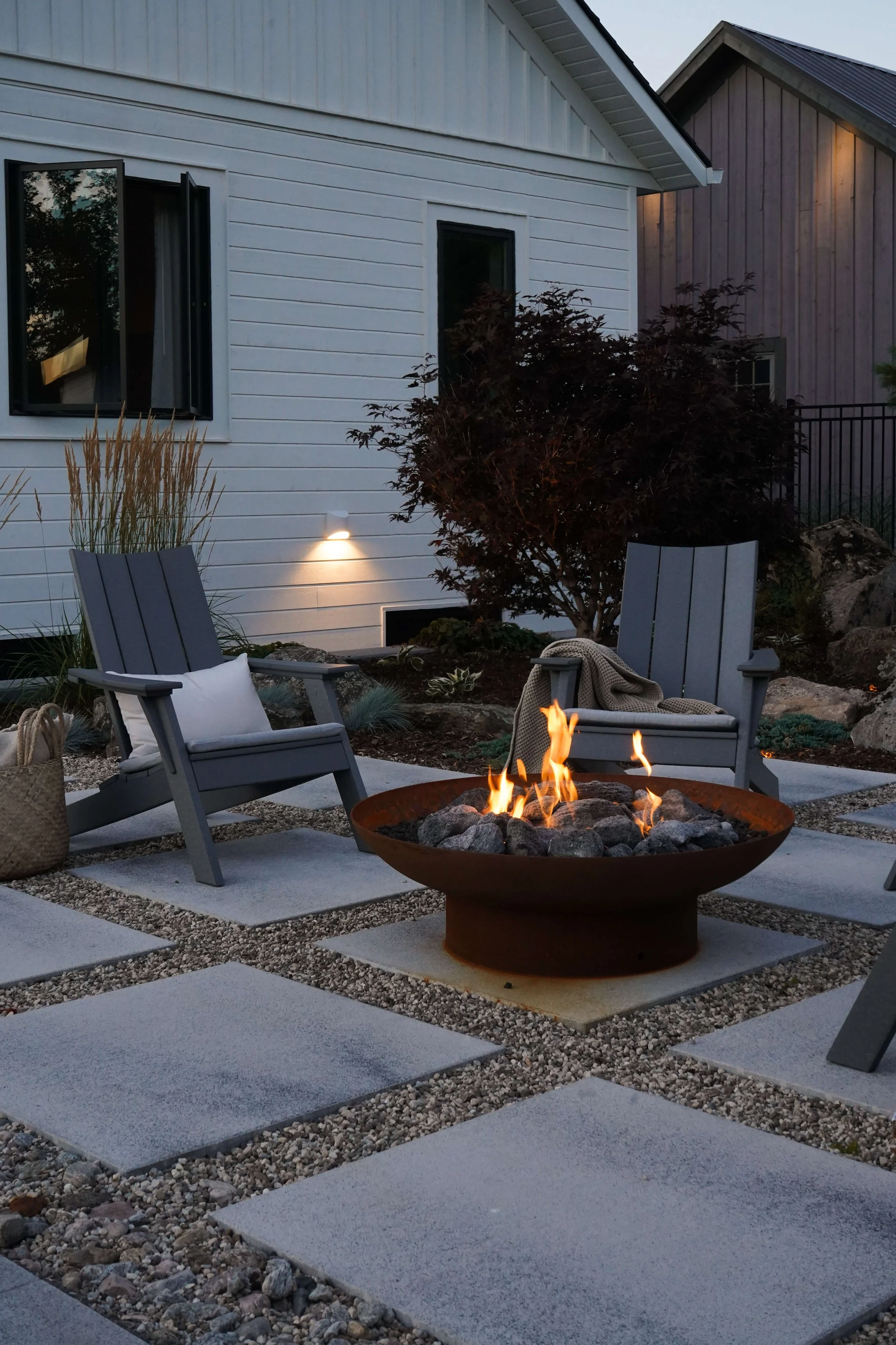 A backyard patio with two gray Adirondack chairs, a fire pit with a flame, potted plants, and surrounding rocks and pavers at dusk.