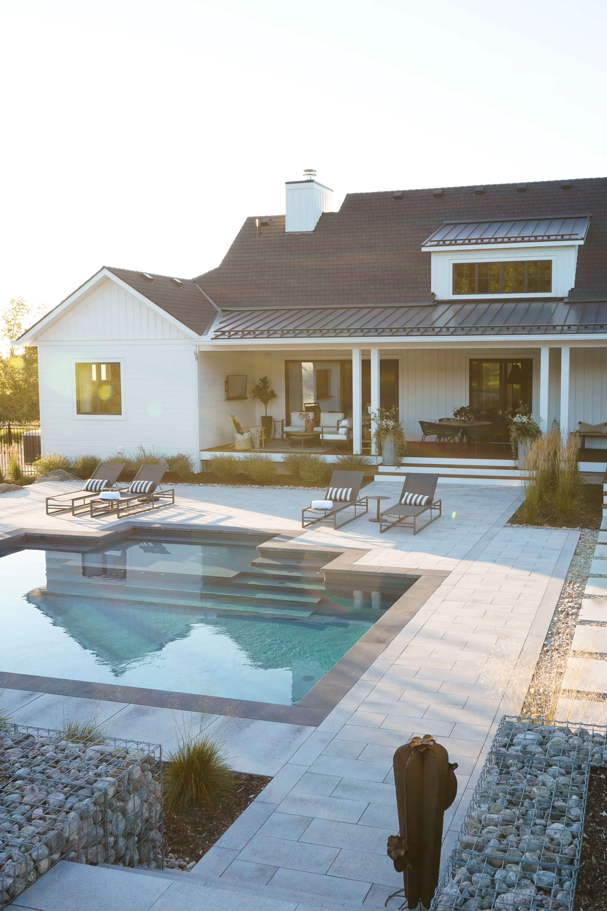 A sunny backyard with a swimming pool, poolside lounge chairs, and a modern house with a large porch.