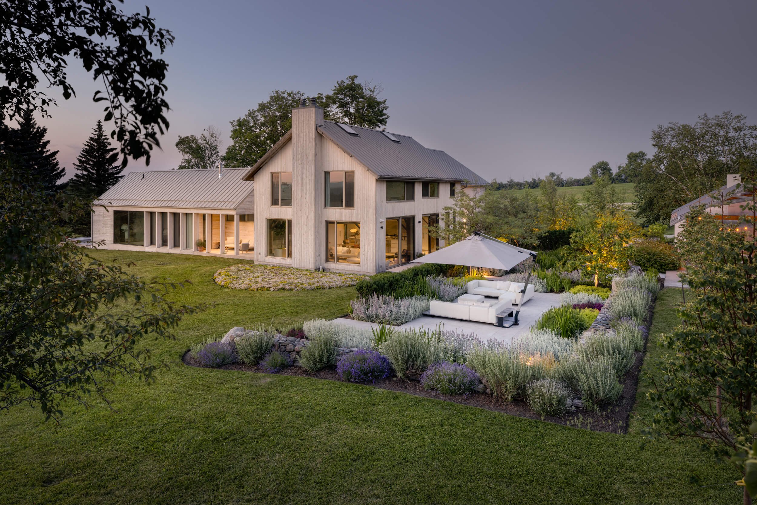 Modern two-story house with large windows, surrounded by a landscaped yard with a patio, outdoor furniture, and garden plants.