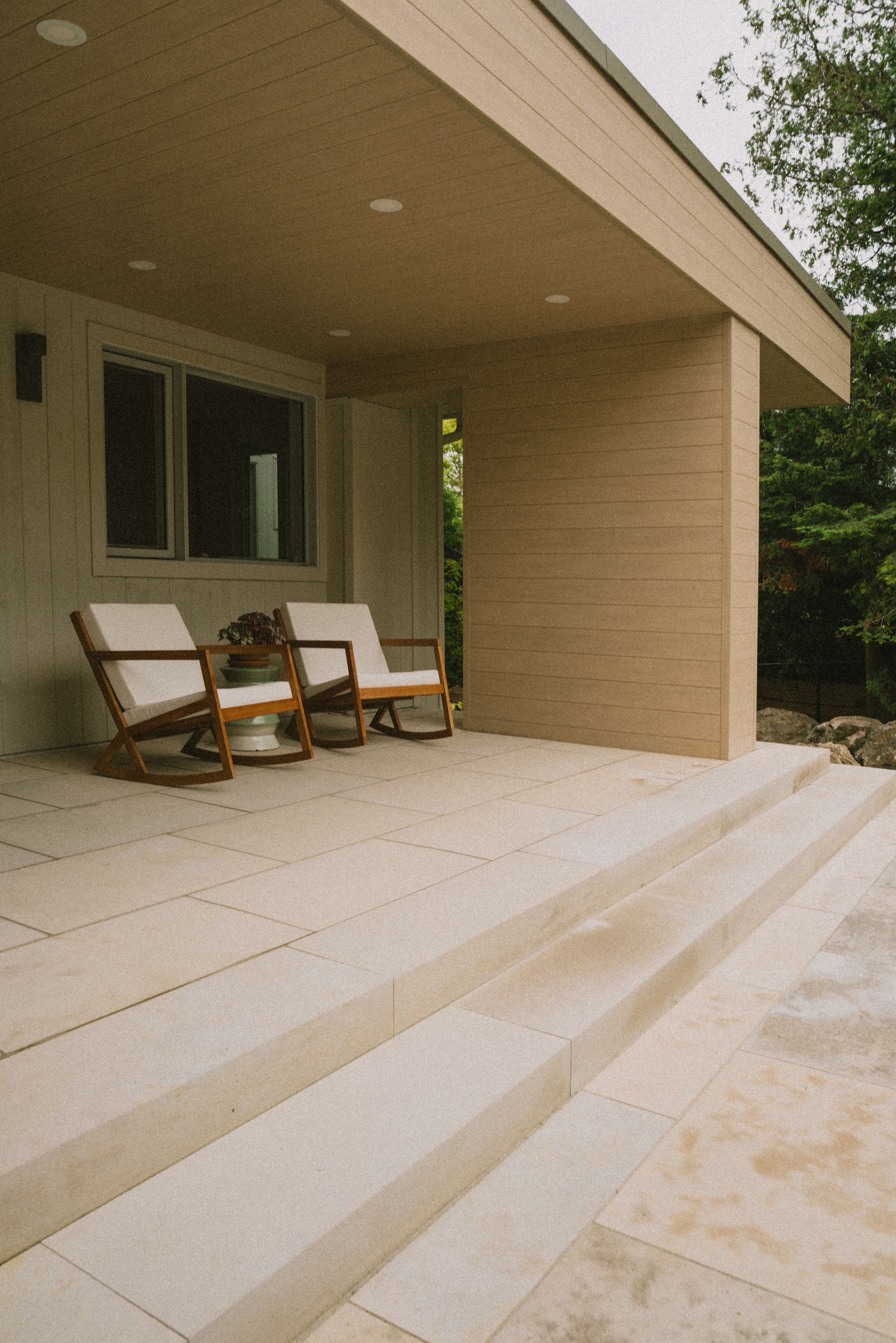 A modern patio with beige stone flooring, three wooden chairs with white cushions, a scenic view of green trees, and a covered roof with recessed lighting.