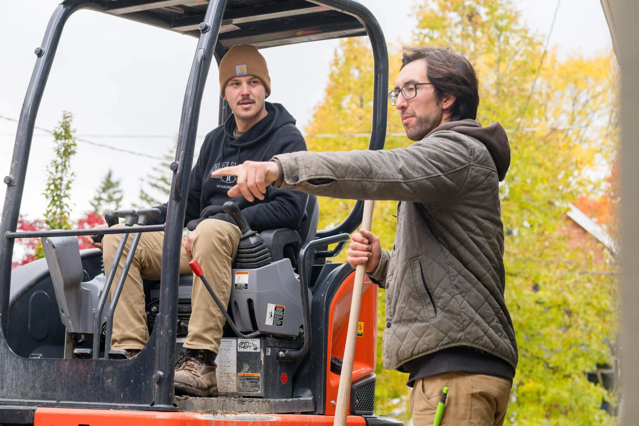 Two men engaged in a discussion outdoors with autumn-colored trees in the background. One man is sitting on a small construction vehicle, dressed in casual work attire, and listening attentively. The other man is standing beside him, gesturing and holding a tool or handle, also dressed in casual work clothes.