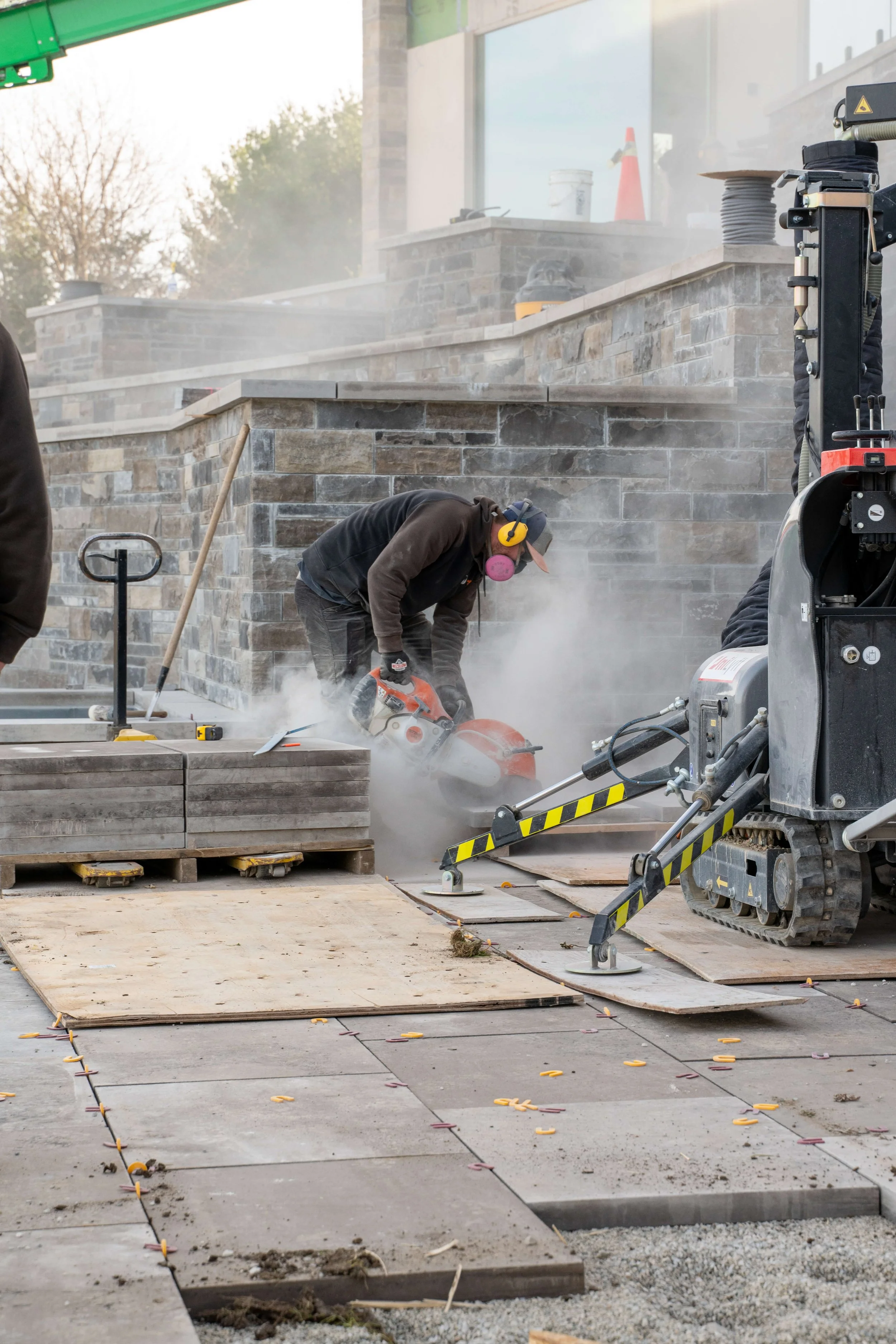 A construction worker using a power saw to cut stone tiles at an outdoor worksite with brick walls, pallets of tiles, and construction equipment visible.
