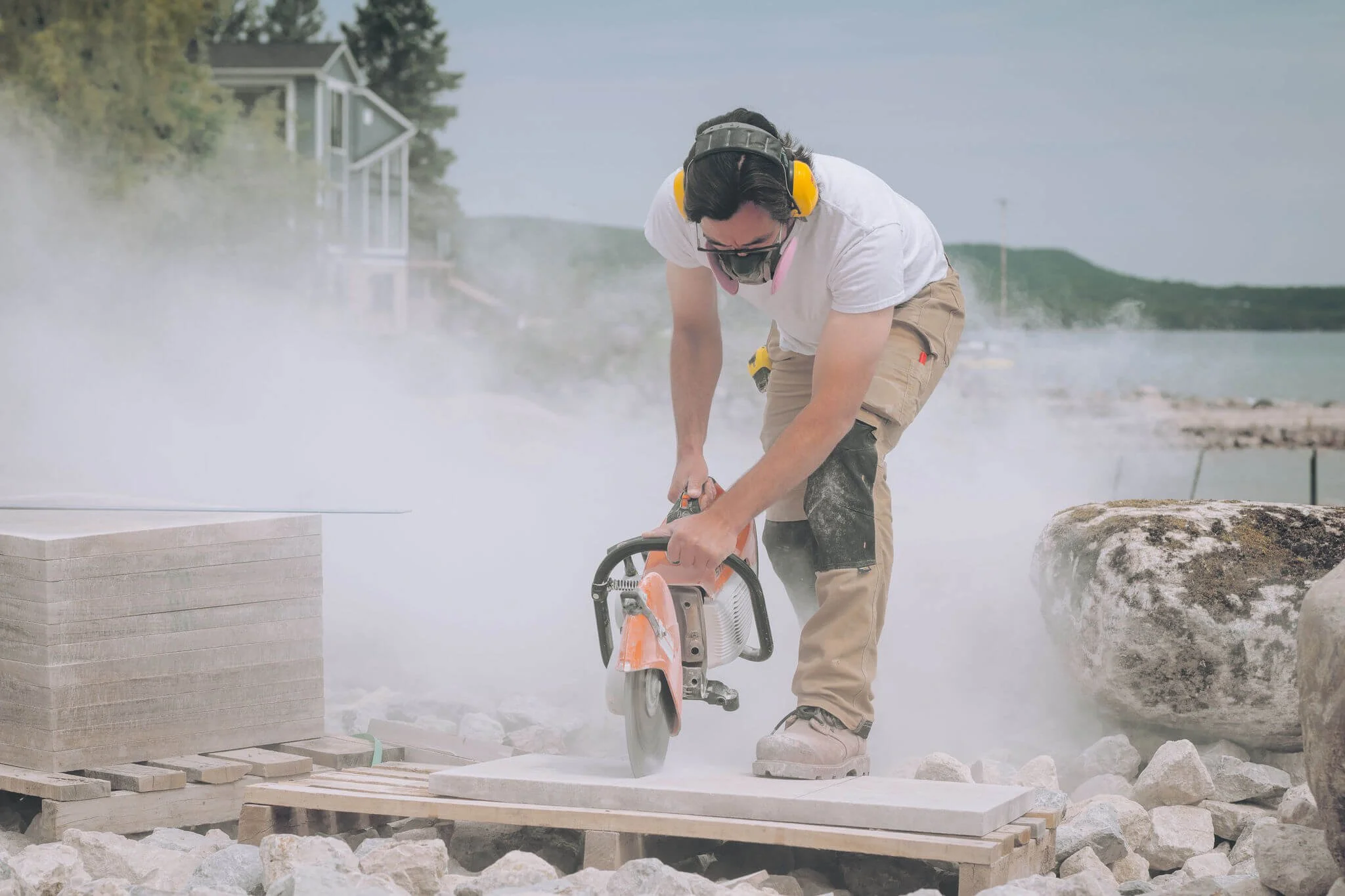A man using a handheld power saw to cut stone outdoors near a lake, with steam or dust rising around him.