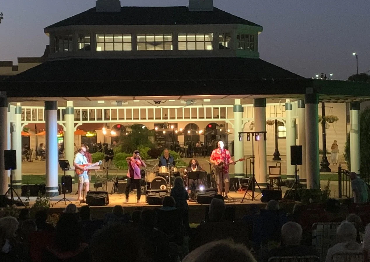 Outdoor concert in Bloomingdale’s Old Town Park bandshell with live music, picnic-goers on the lawn, Mahan Family Wines served, and food from a weekly food truck