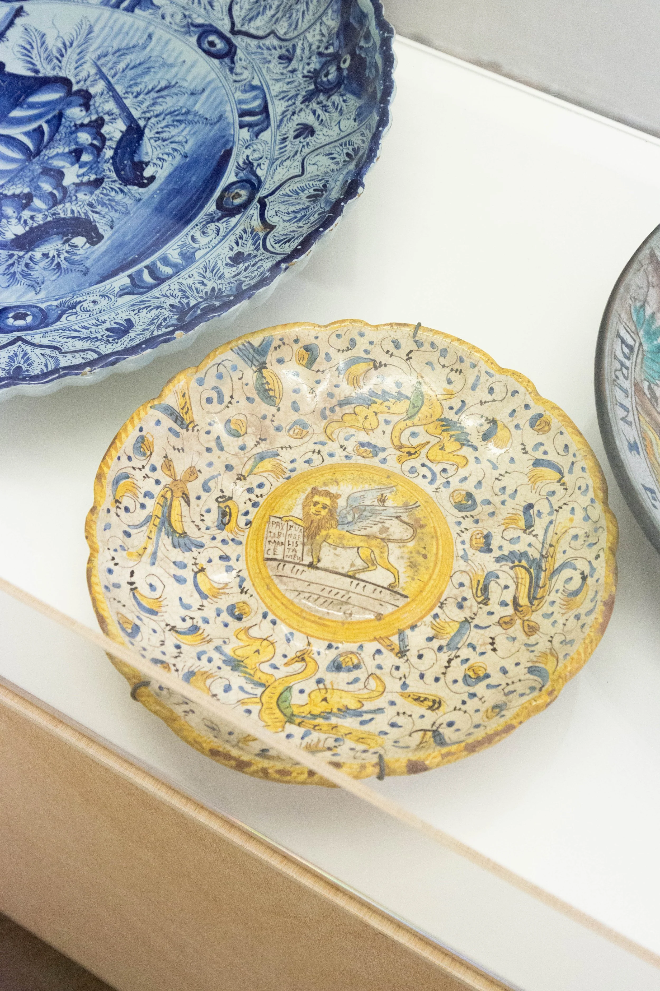   fig37   le monde , 2026, plywood, cardboard, acrylic glass, 86,5 x 125 x 50 cm, Tazza, Italy, 19th century, Majolica, polychrome painted depiction of the Lion of St. Mark with inscription, grotesque ornament. Chipped, with wall, mounting, Ø 24 cm, 