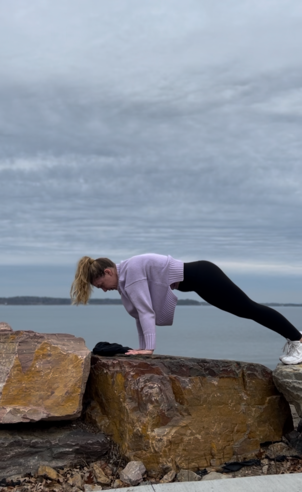 A woman does yoga and exercise by Lake Champlain in black Lululemon tights. She holds a plank.