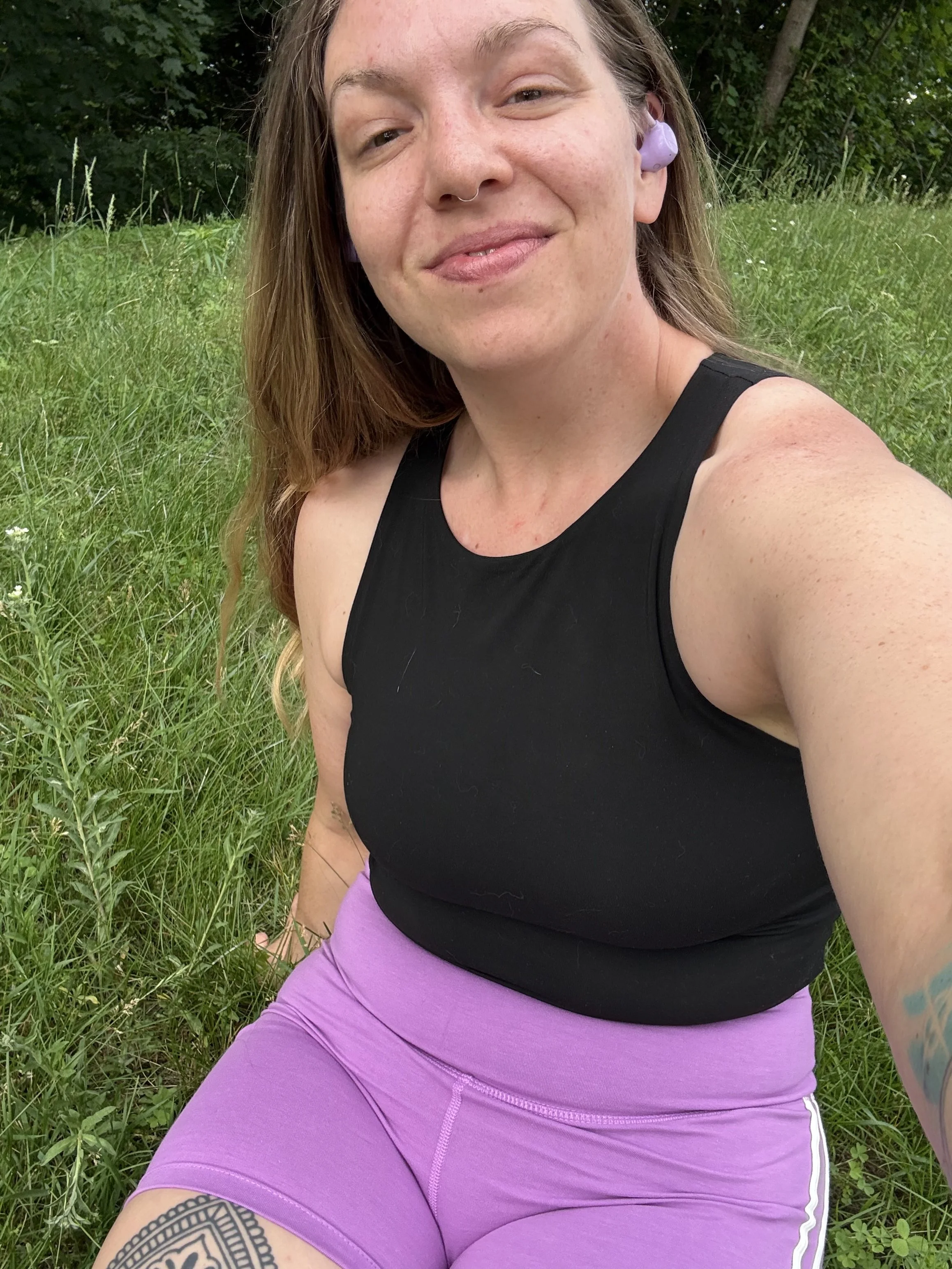 A woman with long hair and a septum piercing smiling outdoors, wearing a black tank top and purple athletic leggings, with greenery in the background.