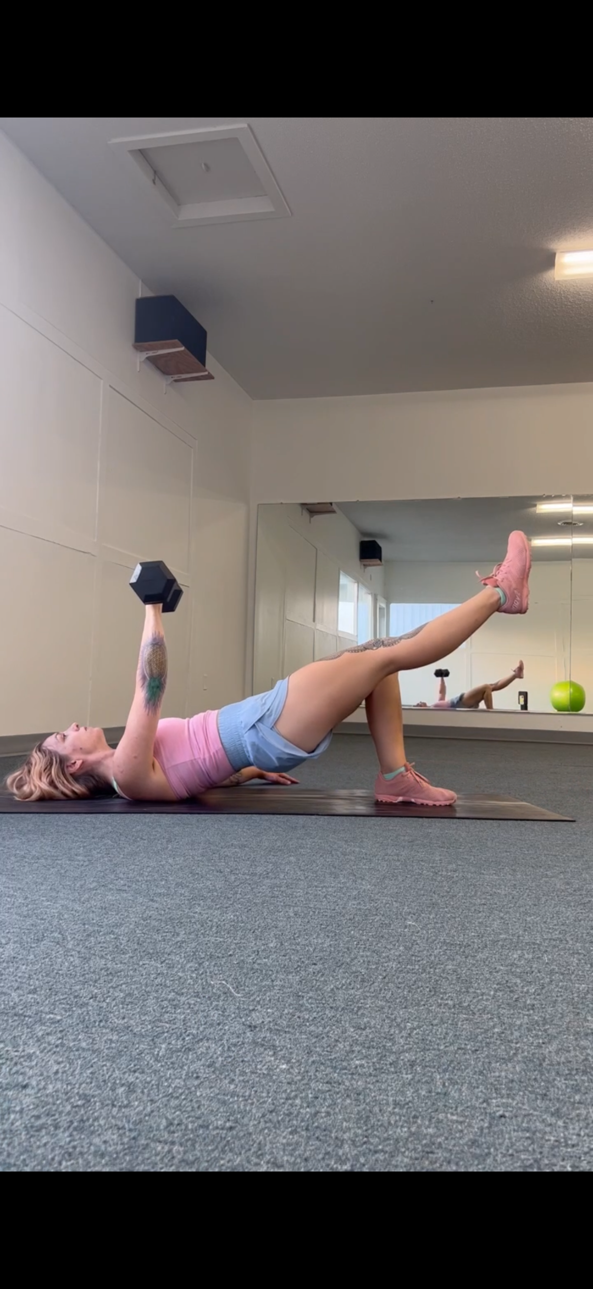 A woman is on her back in a single leg hip raise, while doing a chest press with one dumbbell at the same time