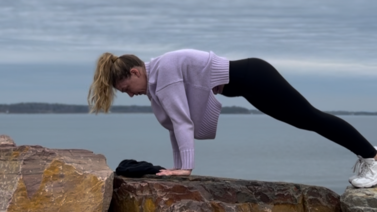 A woman in a purple sweater and black leggings with white shoes performs a plank exercise next to Lake Champlain