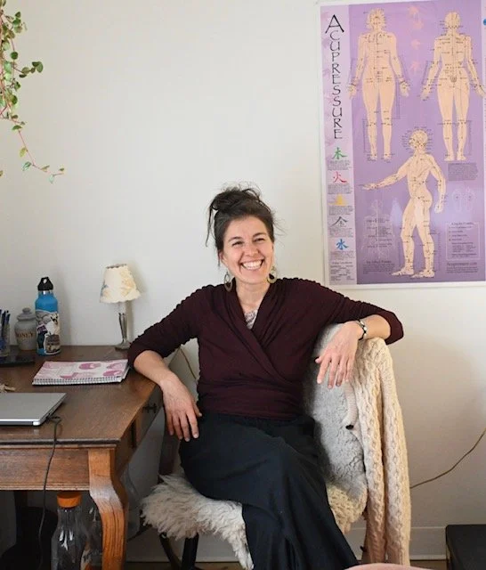 A woman sitting in a cozy office or therapy room, smiling at the camera. She is wearing a dark top and a long dark skirt, with her hair up. Behind her is a wall with a large colorful medical poster, and to her left are a desk, a computer, a water bottle, and a small lamp.
