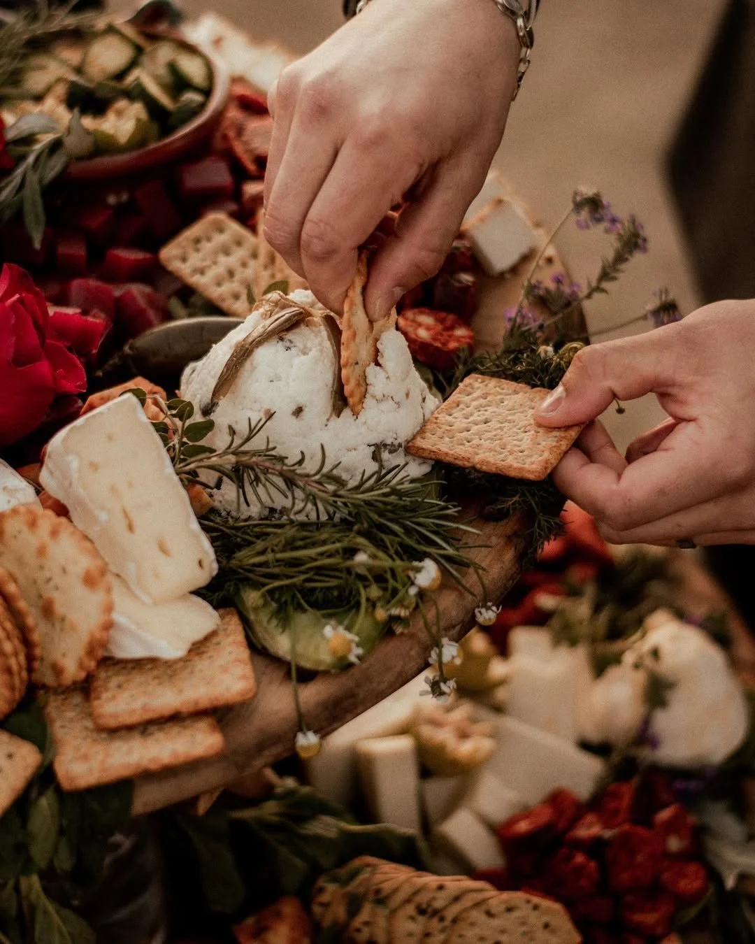Hands reaching for crackers and arranging a cheese and charcuterie board with various cheeses, crackers, herbs, and flowers.
