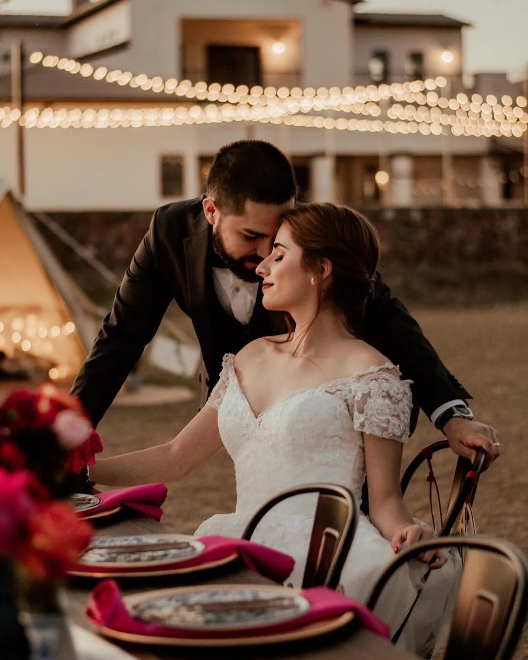 A bride and groom sharing an intimate moment at an outdoor wedding reception during sunset, with string lights and a building in the background.