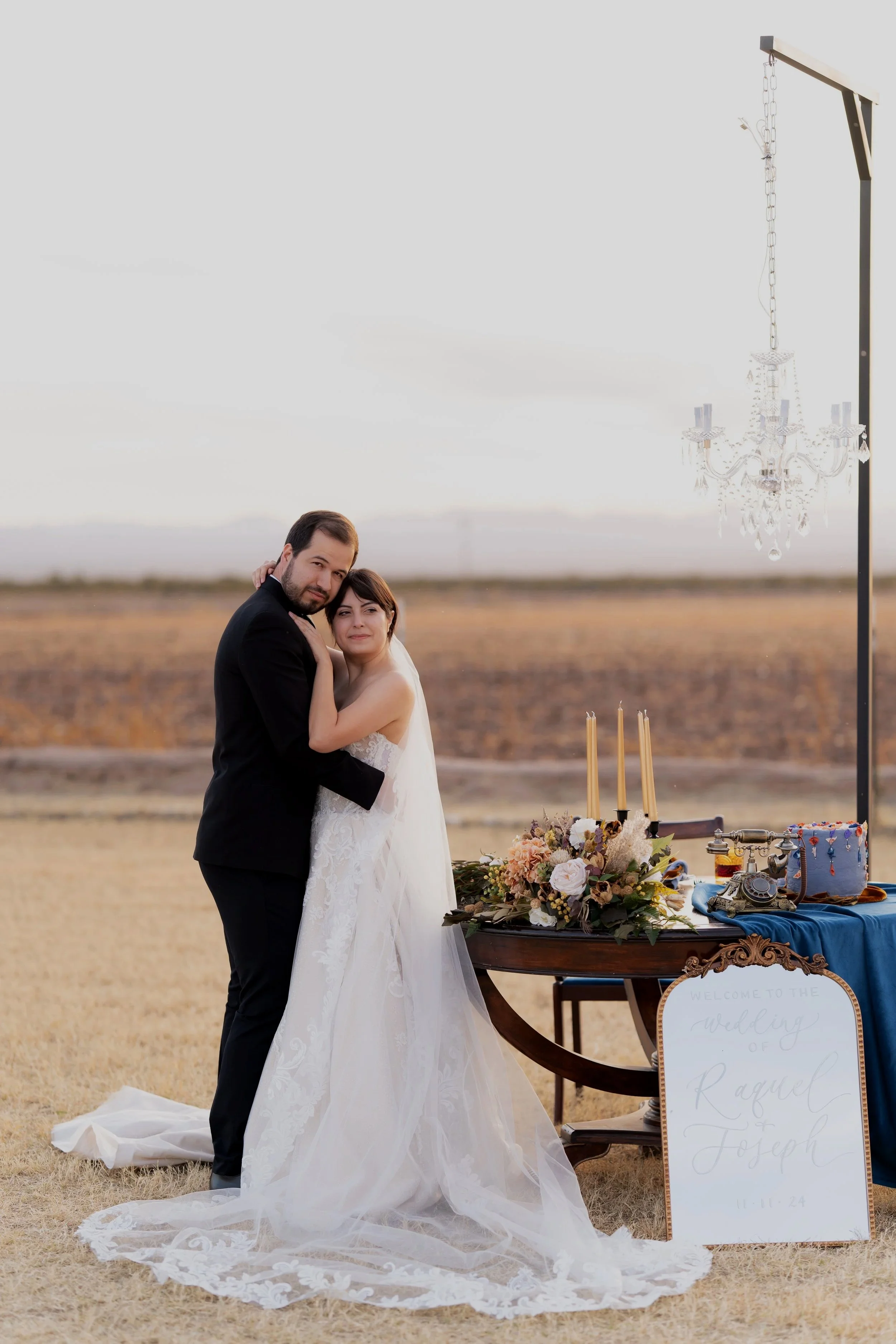A bride and groom hugging outdoors during sunset on their wedding day, with a table of floral arrangements, candles, and vintage decor next to them, and a chandelier hanging from a metal frame.