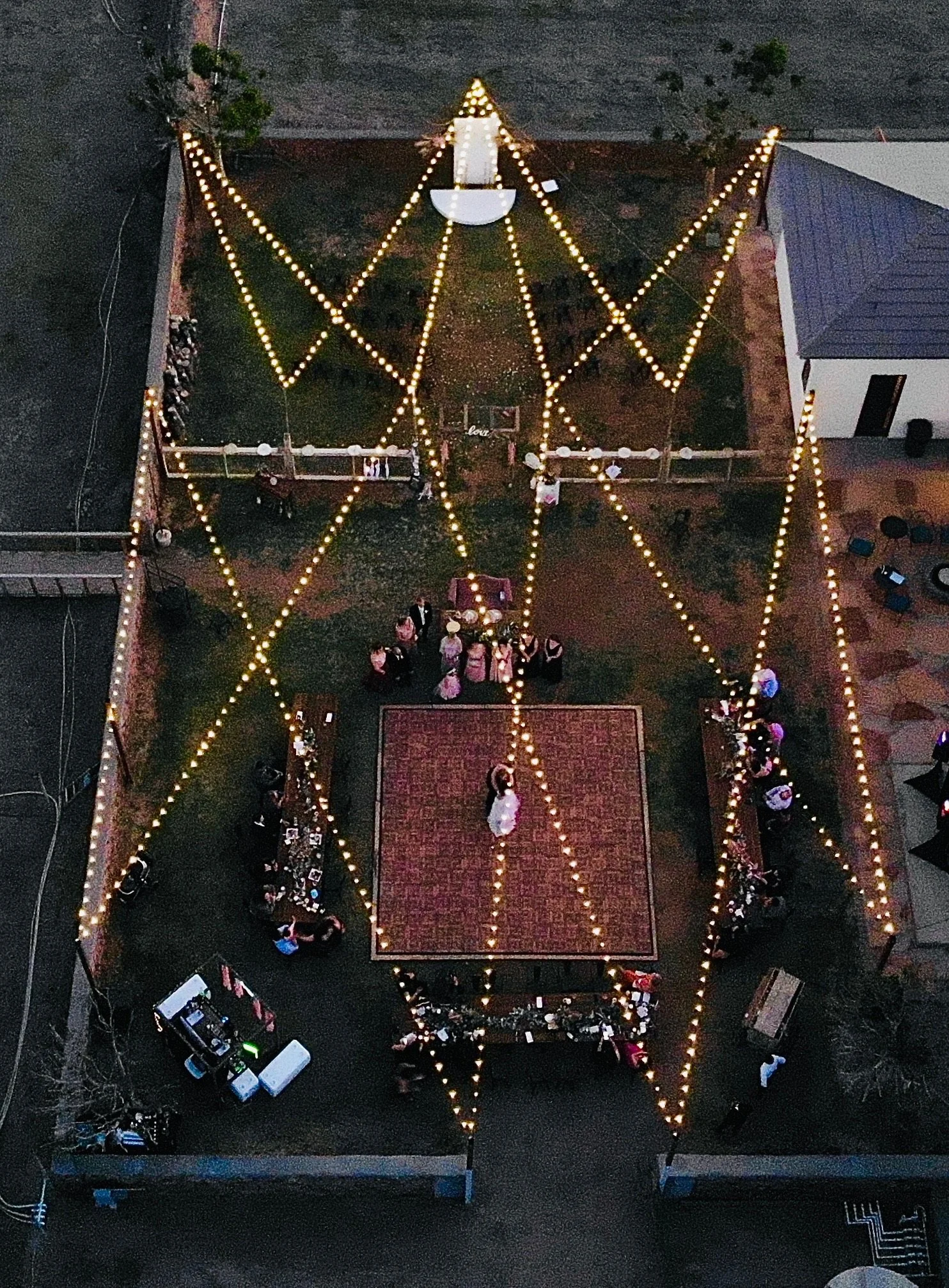 An aerial view of an outdoor wedding reception area decorated with string lights, a dance floor, and tables for guests. The bride and groom are dancing in the center.