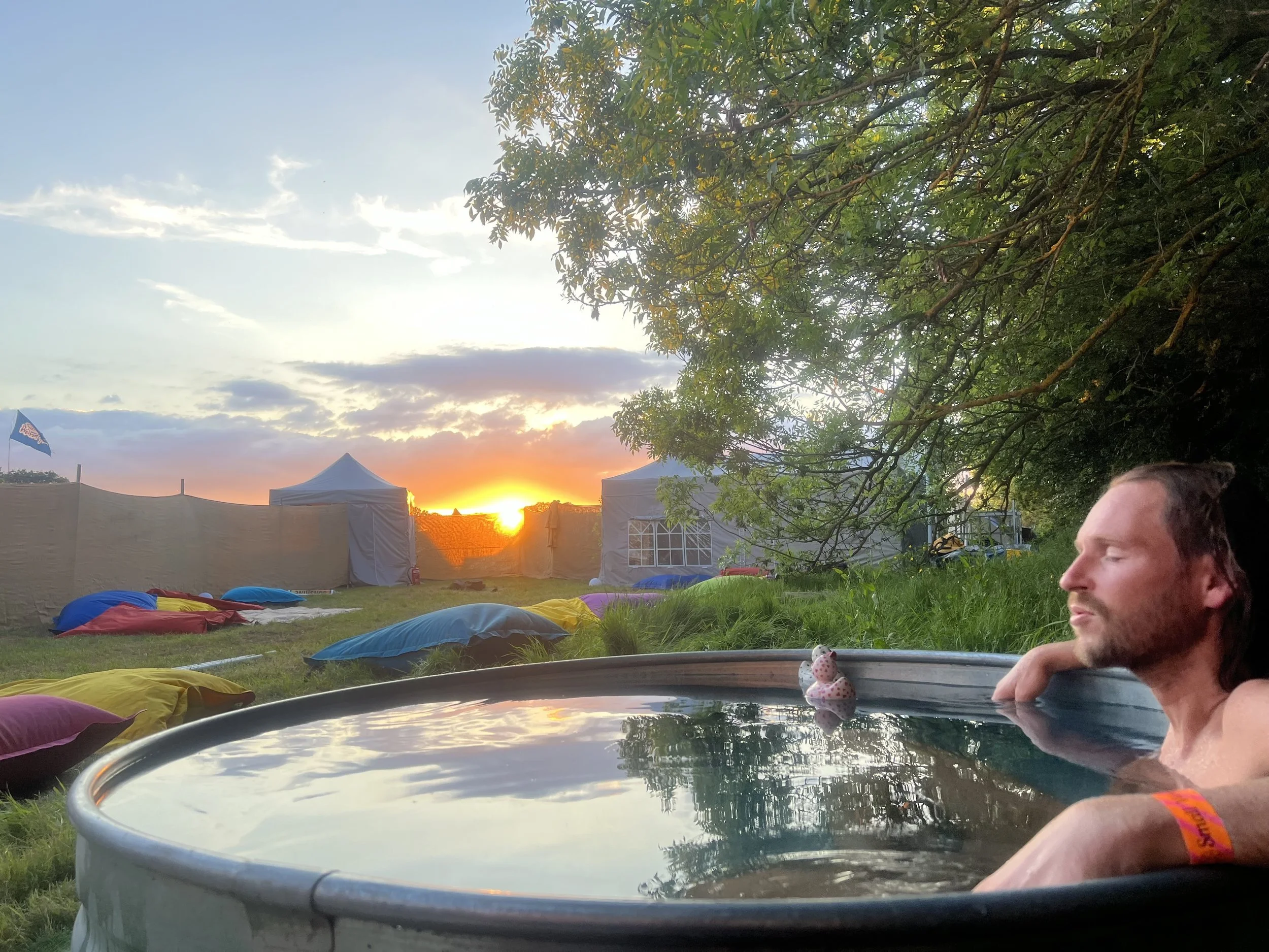A man relaxes in an outdoor hot tub during sunset, with colorful bean bags scattered on the grass nearby and tents set up in the background under a tree.