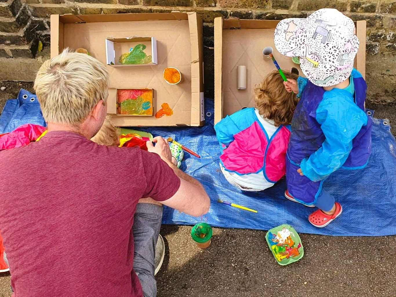 Man and children painting and playing with toys outside on a blue tarp. The kids are dressed in colorful raincoats and a hat, with boxes and containers of paint and toys around them.