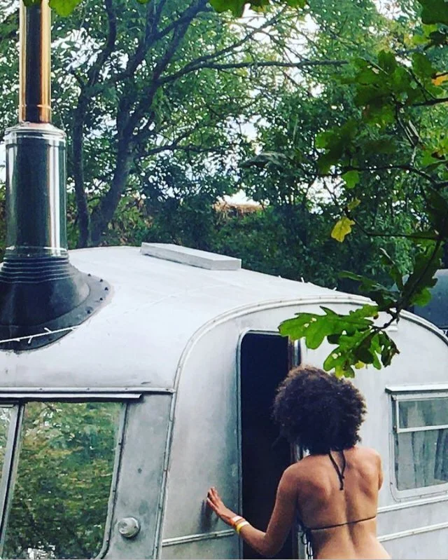 A person with curly hair wearing a black bikini top standing at the entrance of a vintage camper trailer surrounded by trees.