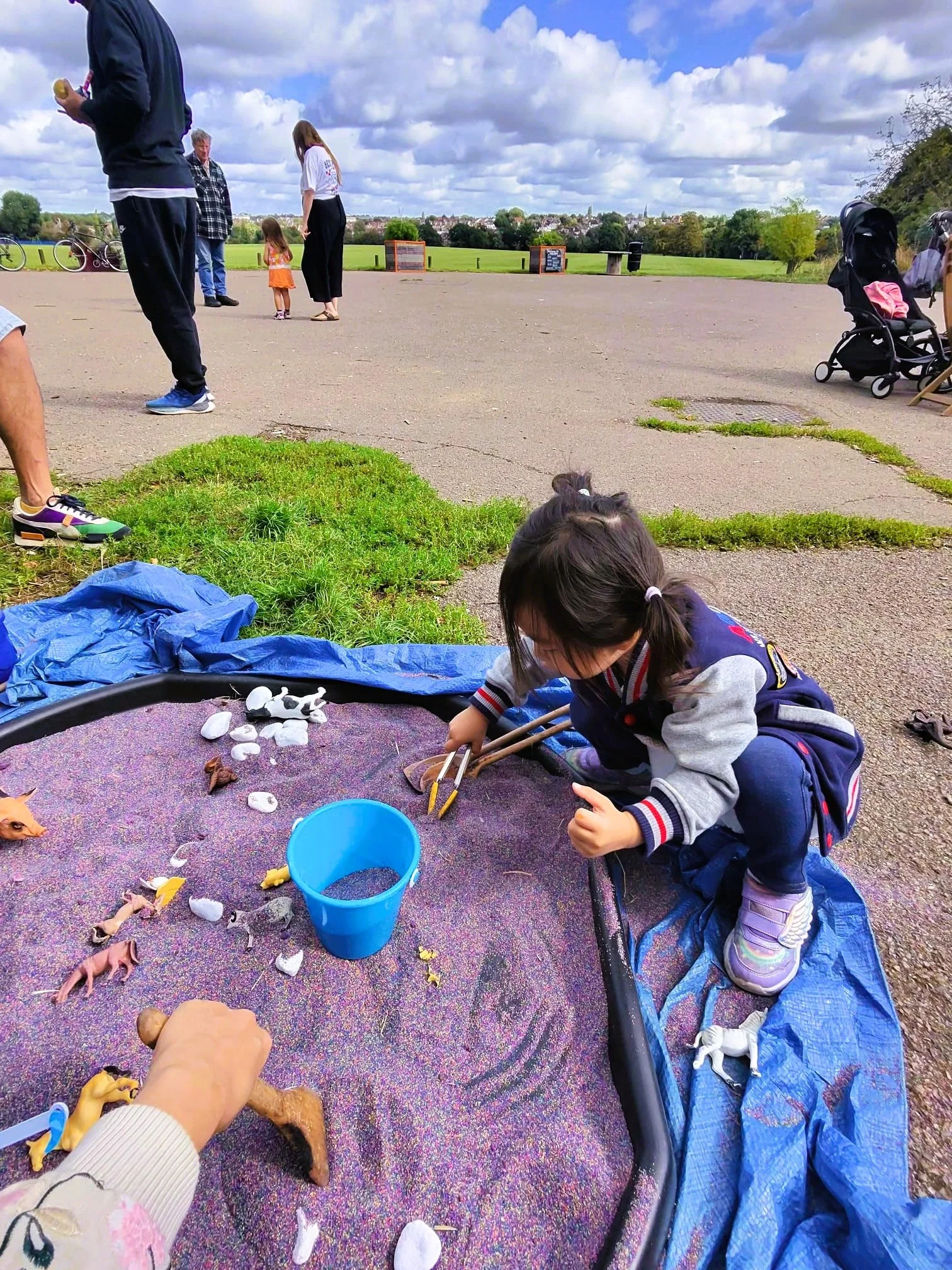 A young girl playing with toy animals at an outdoor sandbox, with people and a stroller in the background on a cloudy day in a park.