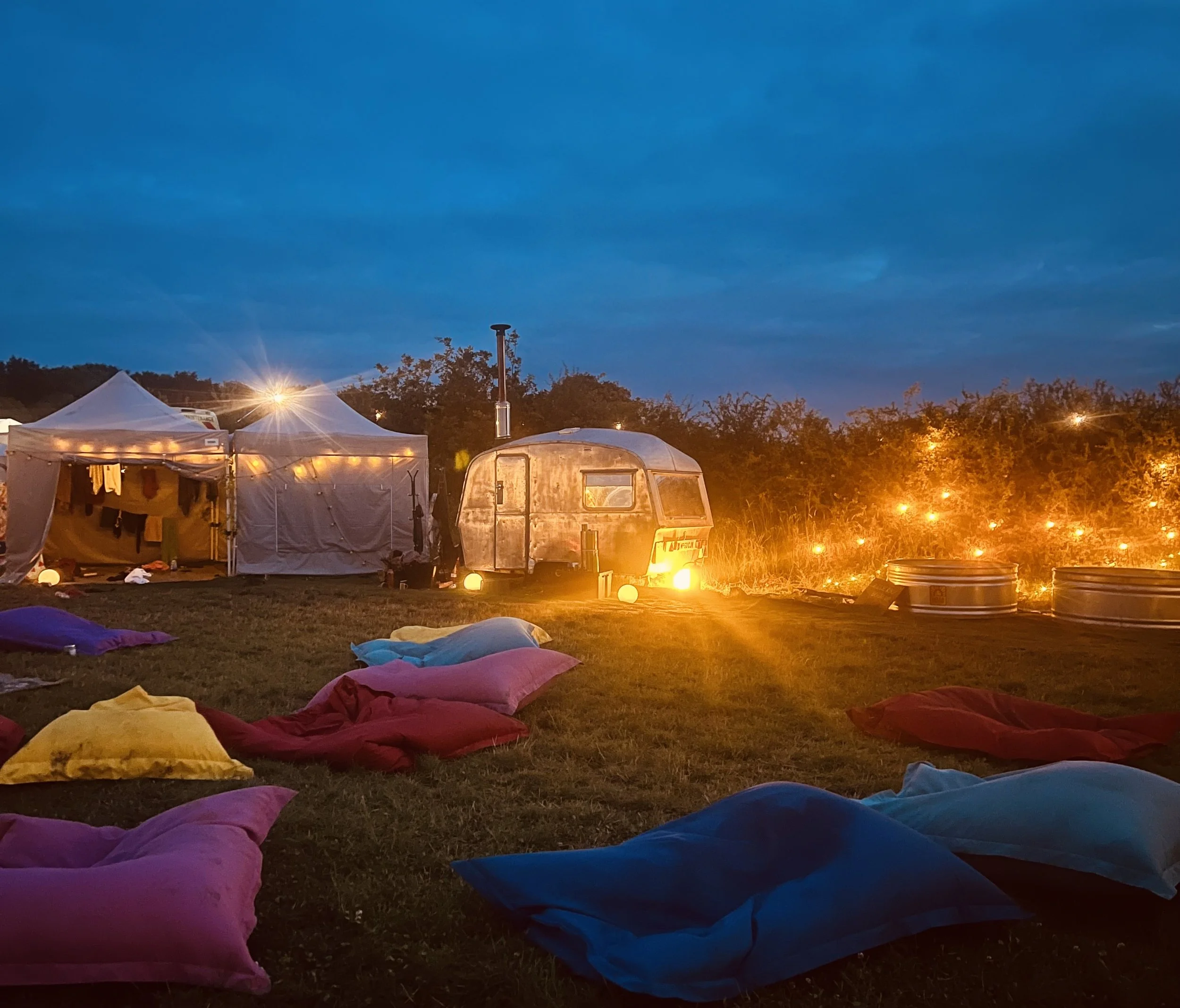 An outdoor camping scene at dusk featuring a small vintage camper trailer, two white tents, colorful sleeping bags and pillows on the ground, and string lights illuminating the area.