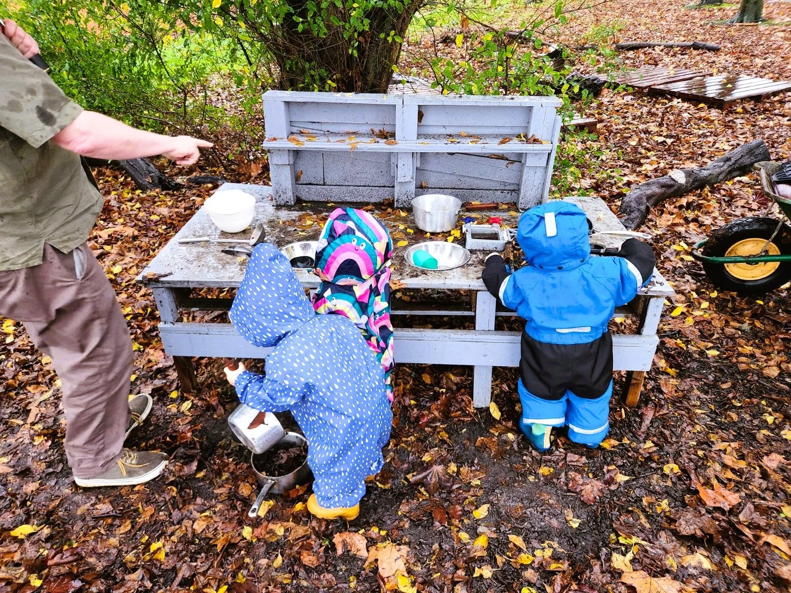 Three children dressed in rain gear playing at a muddy outdoor play kitchen set with a man nearby, surrounded by fallen autumn leaves.