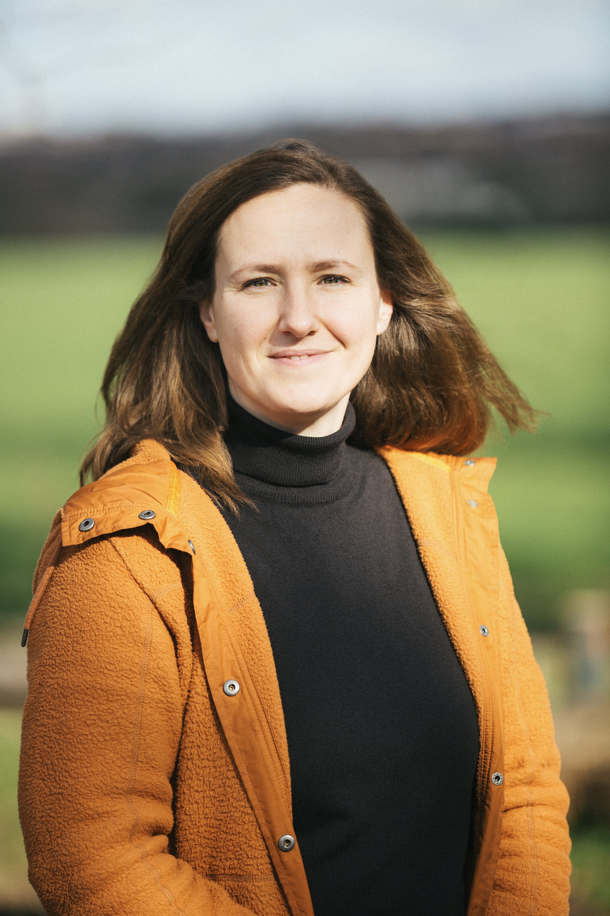 A woman with long brown hair wearing an orange jacket and black turtleneck standing outdoors with a blurred green landscape background.