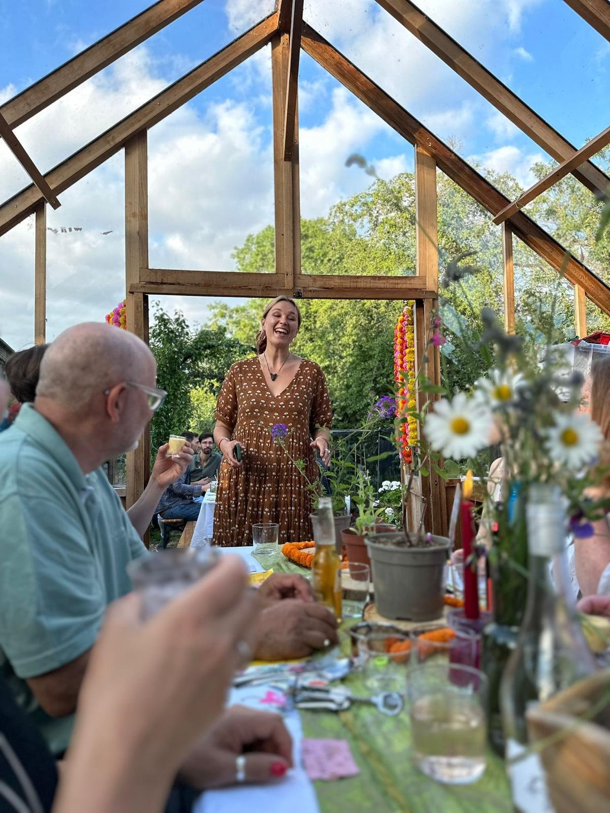 A woman in a brown dress stands smiling and speaking at a dining table marked with conversation, surrounded by guests in a partially built wooden structure outdoors with green trees and a blue sky in the background.