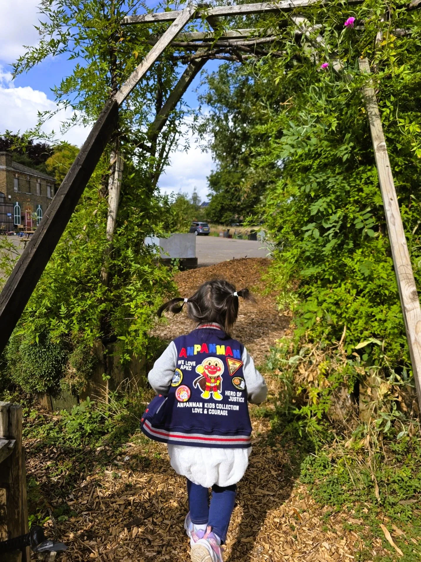 Child with dark hair in pigtails walking under a wooden archway covered in green vines, wearing a jacket with colorful patches, a white dress, and blue tights.