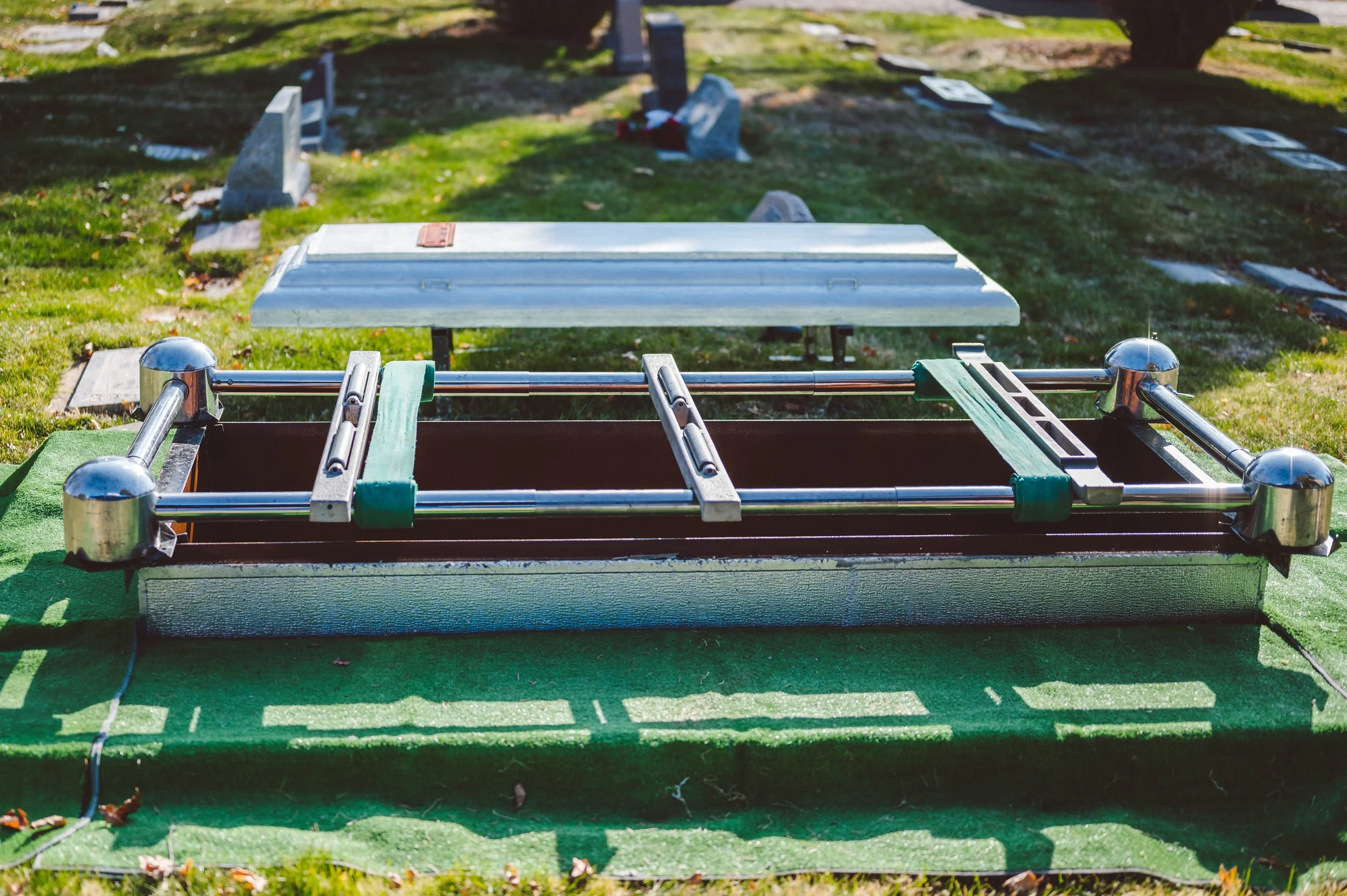 A memorial outdoor setup with a metallic and brown container, possibly a cremation or storage area, with a metallic frame on top. The setup is outdoors on green grass with a grassy background, a tree, and some headstones in the distance.