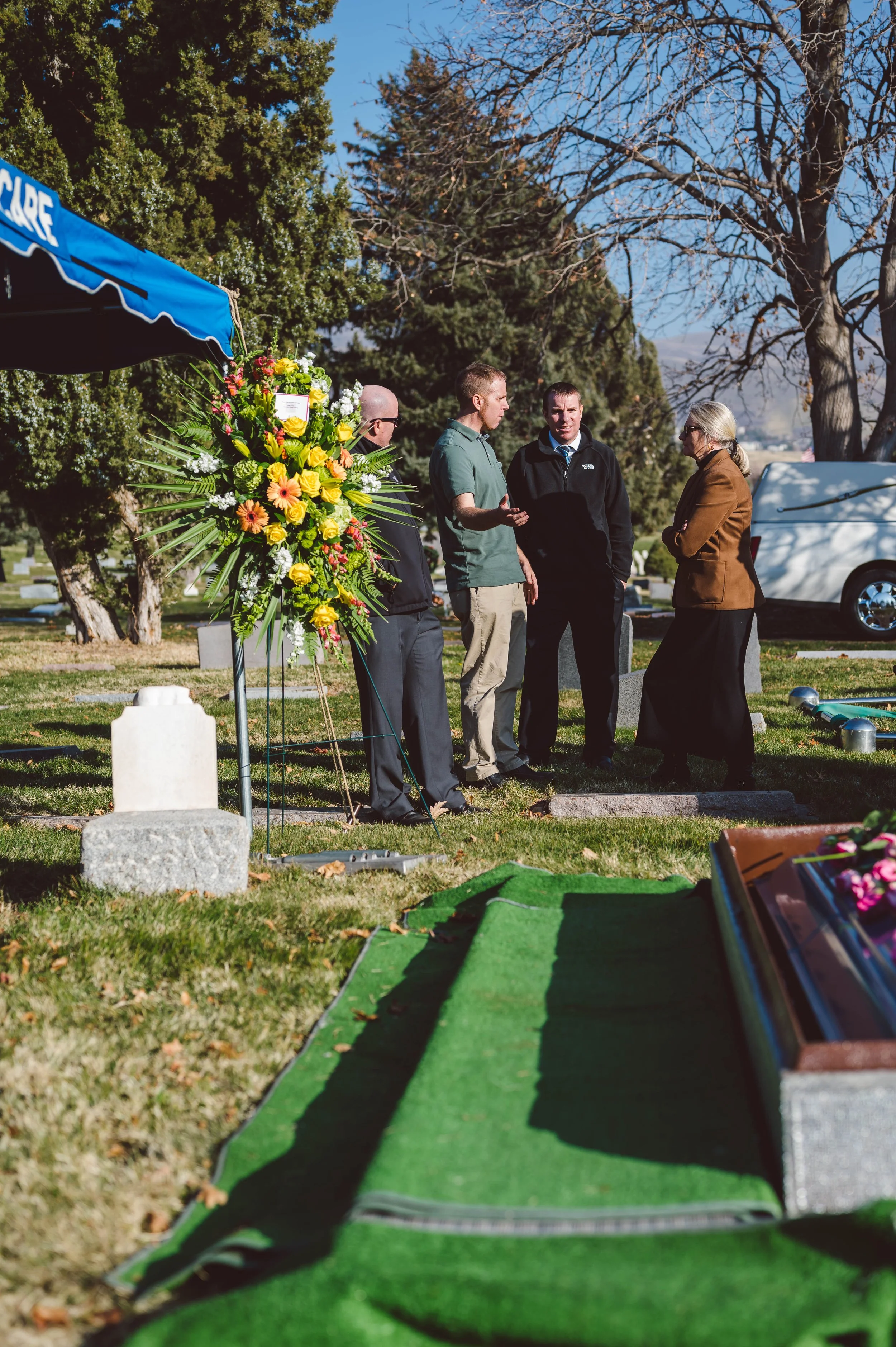 Four people standing and talking at a gravesite during a funeral in a cemetery. There are trees in the background and a large floral arrangement near them.