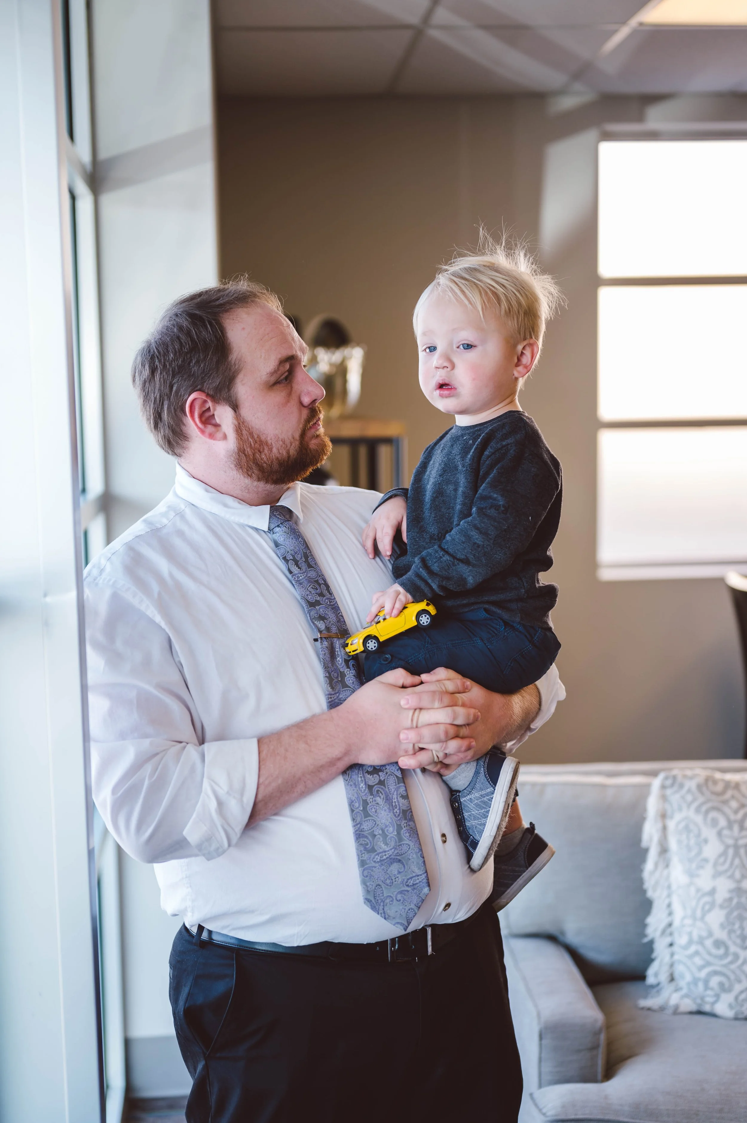 A man with a beard, wearing a white shirt and tie, is holding a young boy with blonde hair, dressed in a dark sweater and pants, indoors with natural light coming through windows in the background.