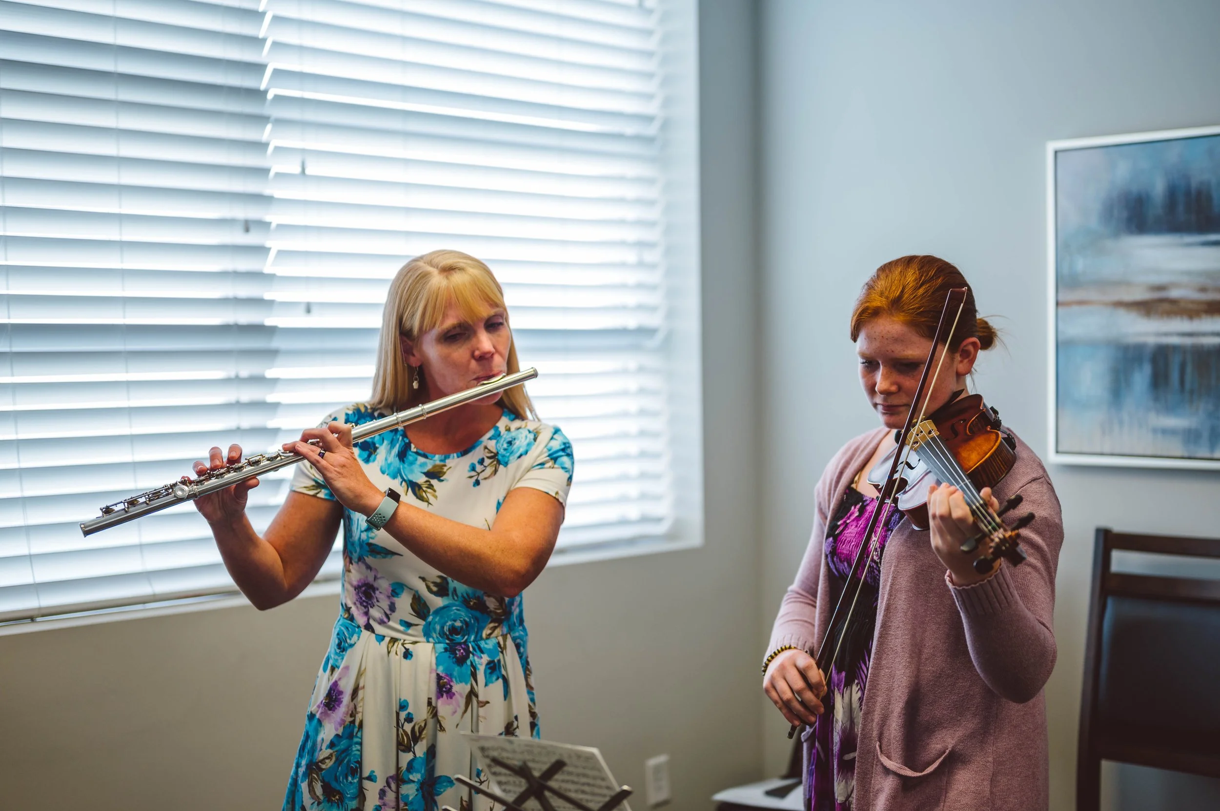 Two women playing musical instruments - a woman playing a flute and another woman playing a violin, indoors near a window with blinds, with a painting on the wall in the background.