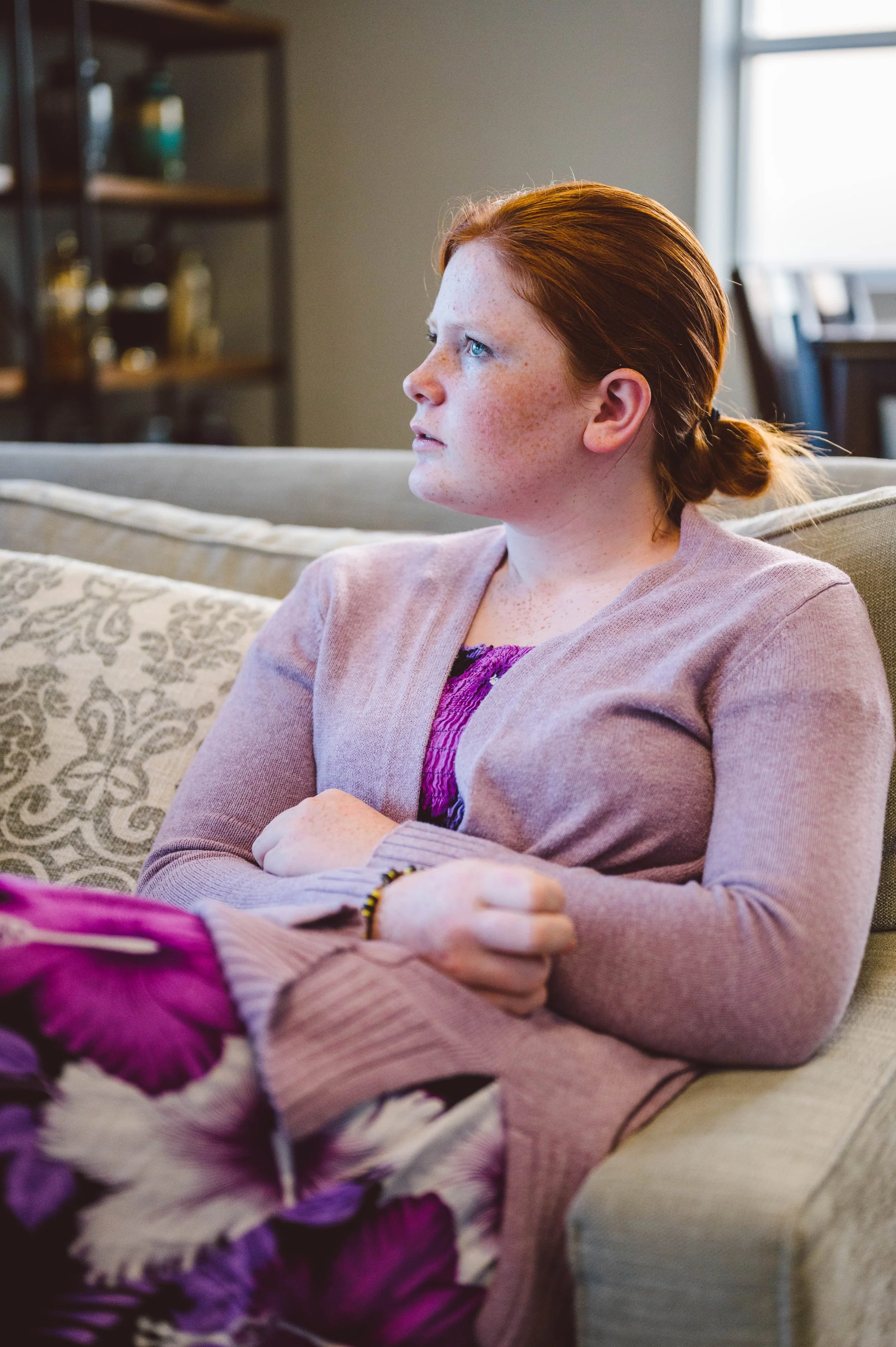 A young woman with red hair and freckles sitting on a beige couch, looking thoughtfully to her left, in a living room with a window and shelves in the background.