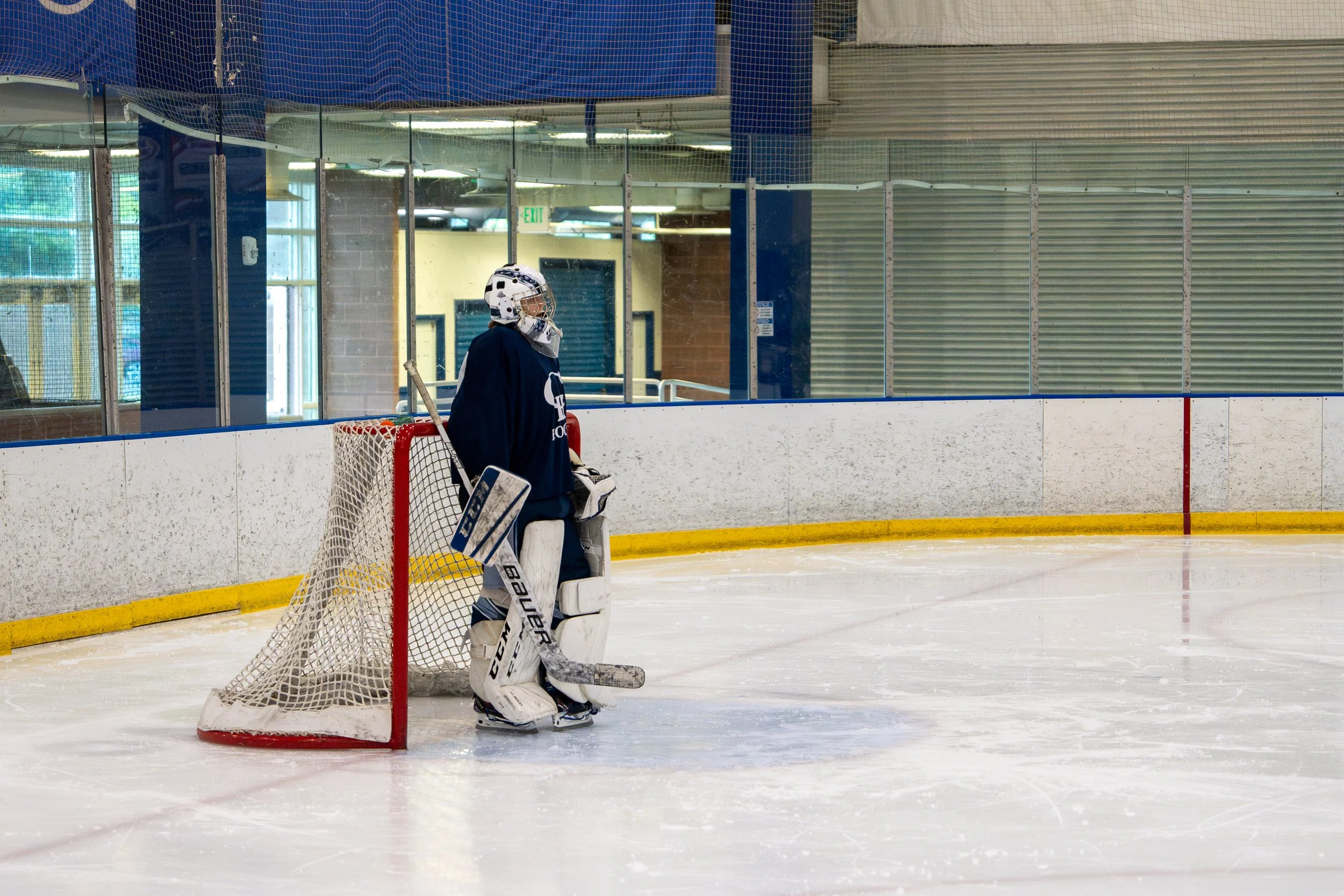 A hockey goalie standing on the ice in an indoor hockey rink, facing away from the camera, wearing full protective gear including a helmet, jersey, and pads, next to the goal net.