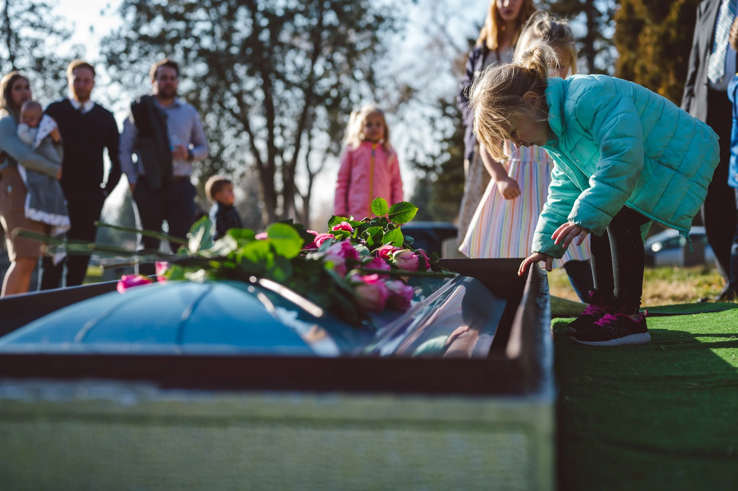 Children and adults gathered outdoors at a grave site, with flowers placed on a coffin or grave, during a memorial or funeral service.