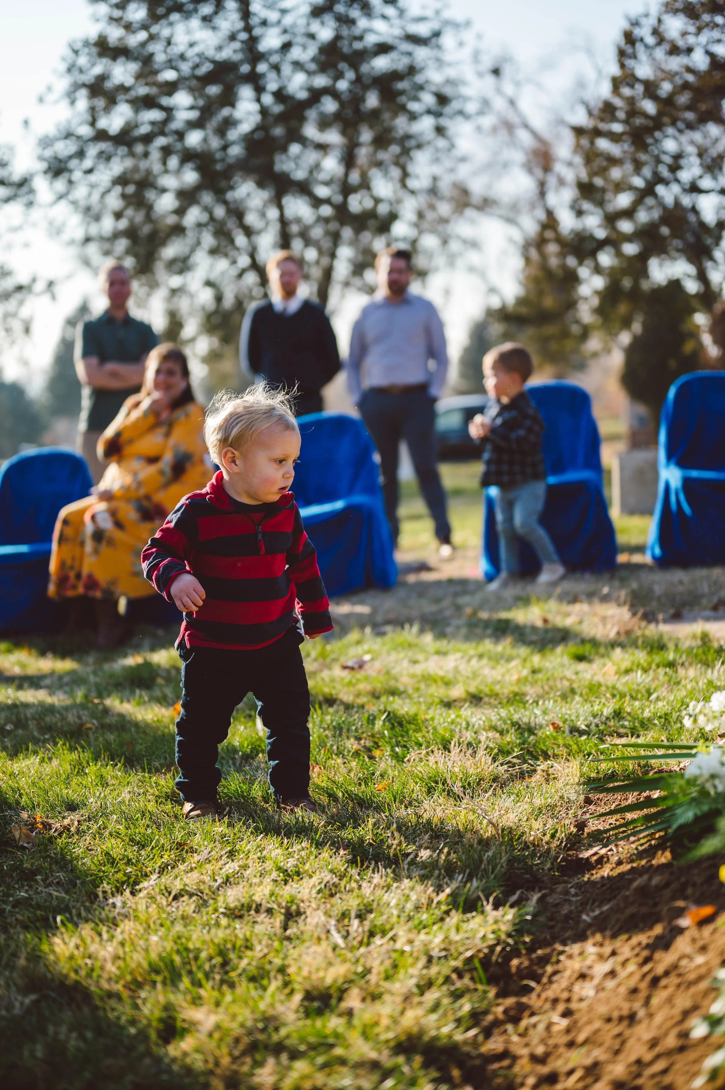 A young boy with blond hair wearing a red and black striped shirt and dark pants, standing outdoors on a grassy area during a daytime gathering with people, including children and adults, in the background.