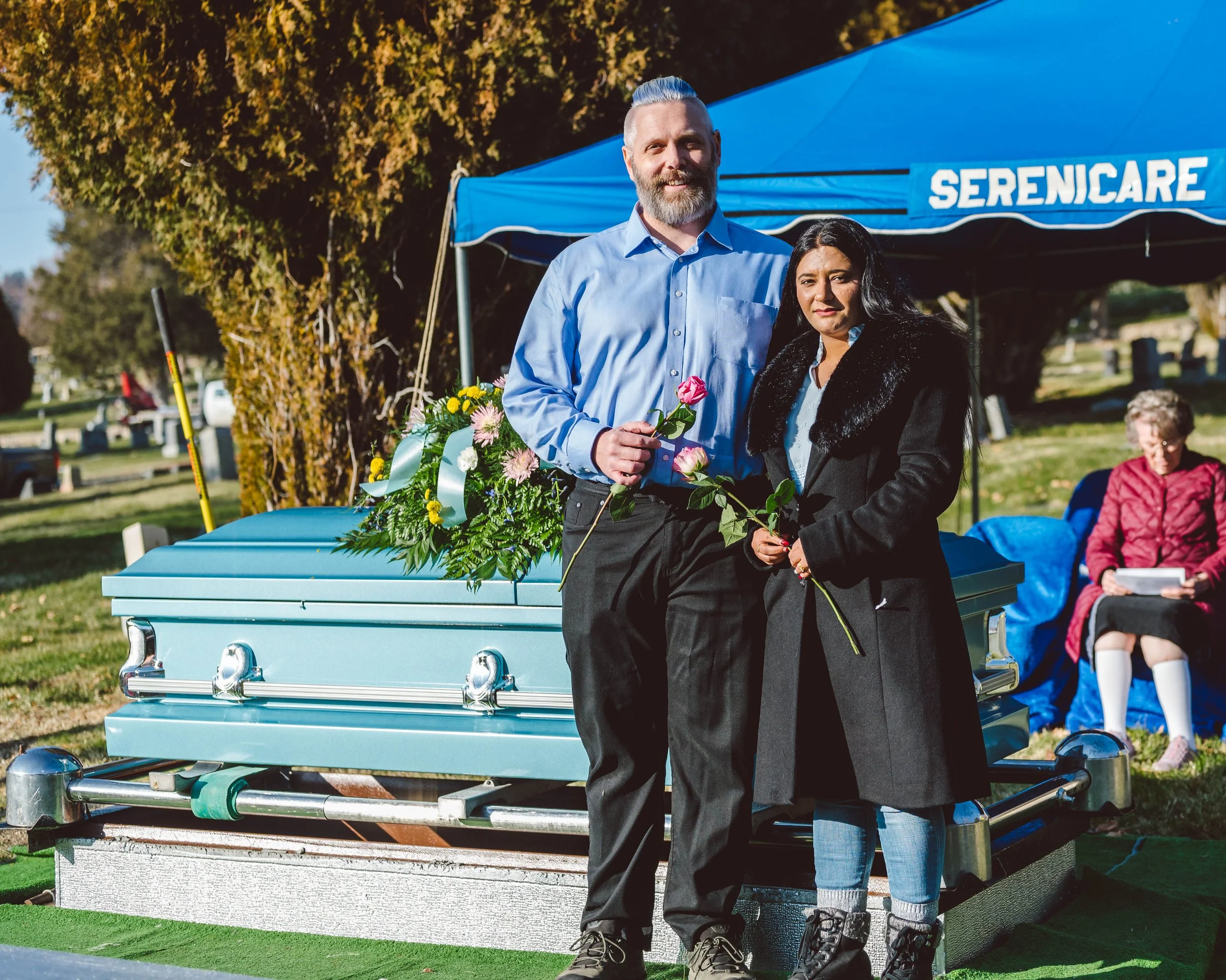 Two people holding flowers standing in front of a blue funeral casket and a cemetery scene.