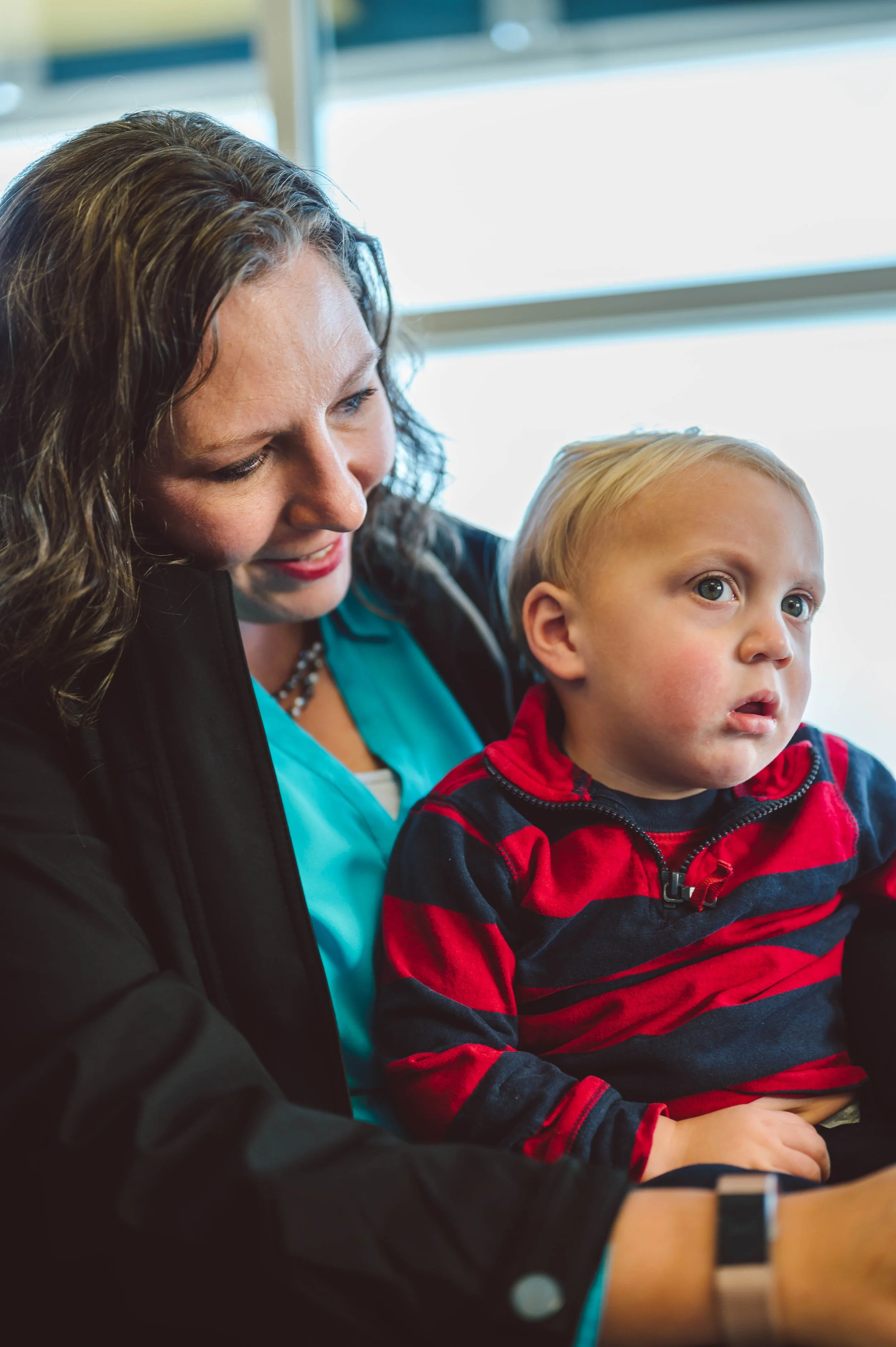 A woman with curly brown hair and wearing a black jacket is holding a young blonde boy in a red and navy striped hoodie, sitting near a window.