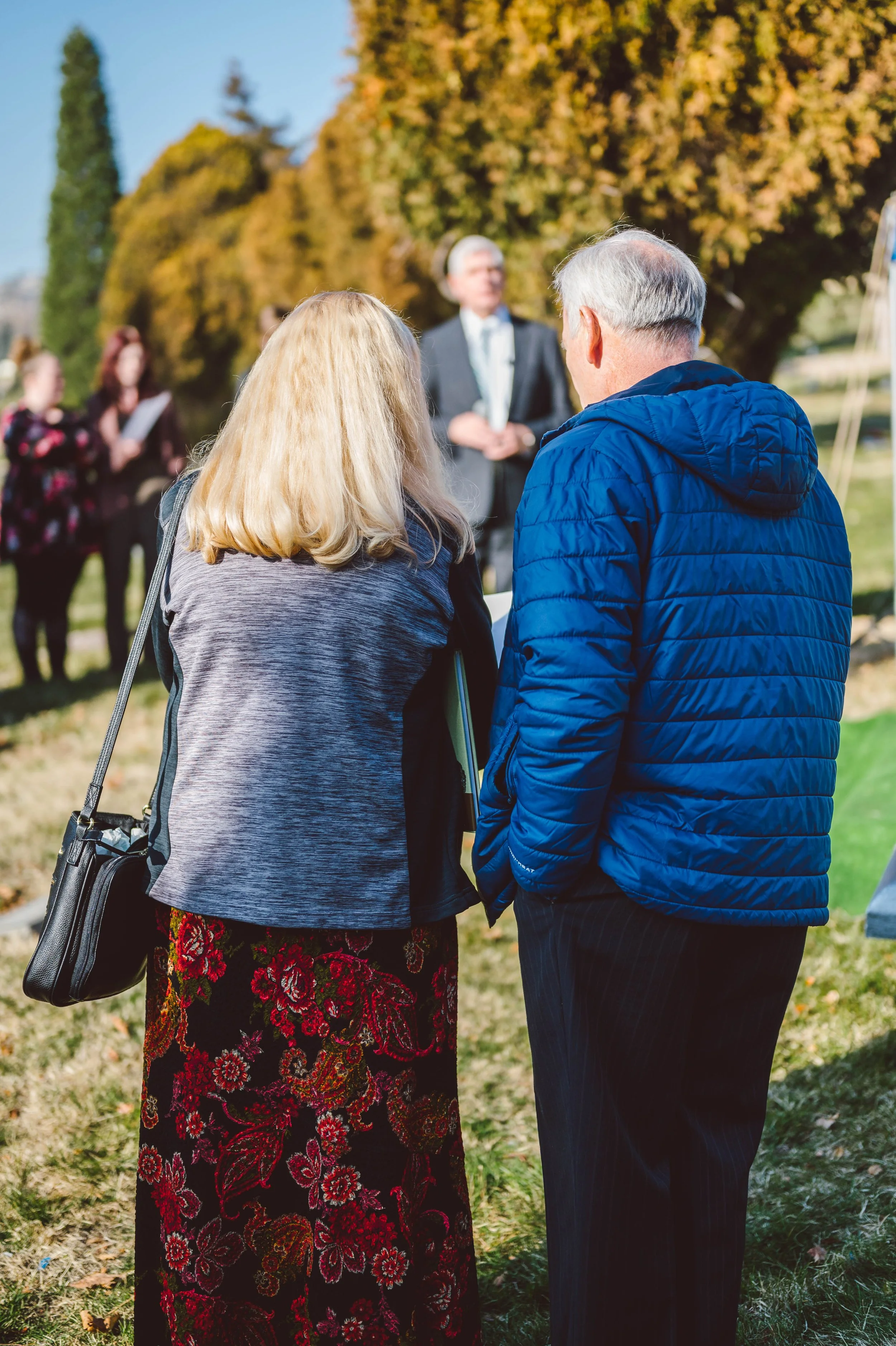 A man and woman standing outdoors during what appears to be a ceremony or gathering, facing a speaker. Trees with autumn foliage are in the background.