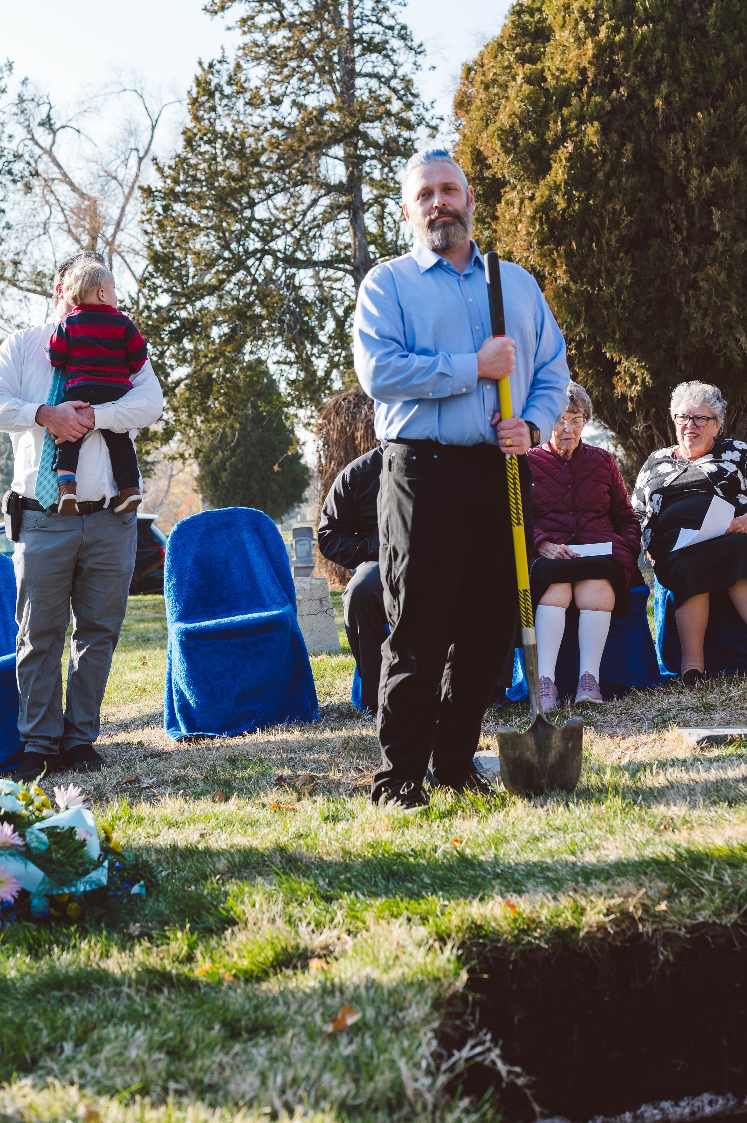 A man with a beard and mustache stands holding a shovel at a gravesite, surrounded by elderly women seated on chairs, and a person holding a child, in a grassy outdoor area with trees in the background.
